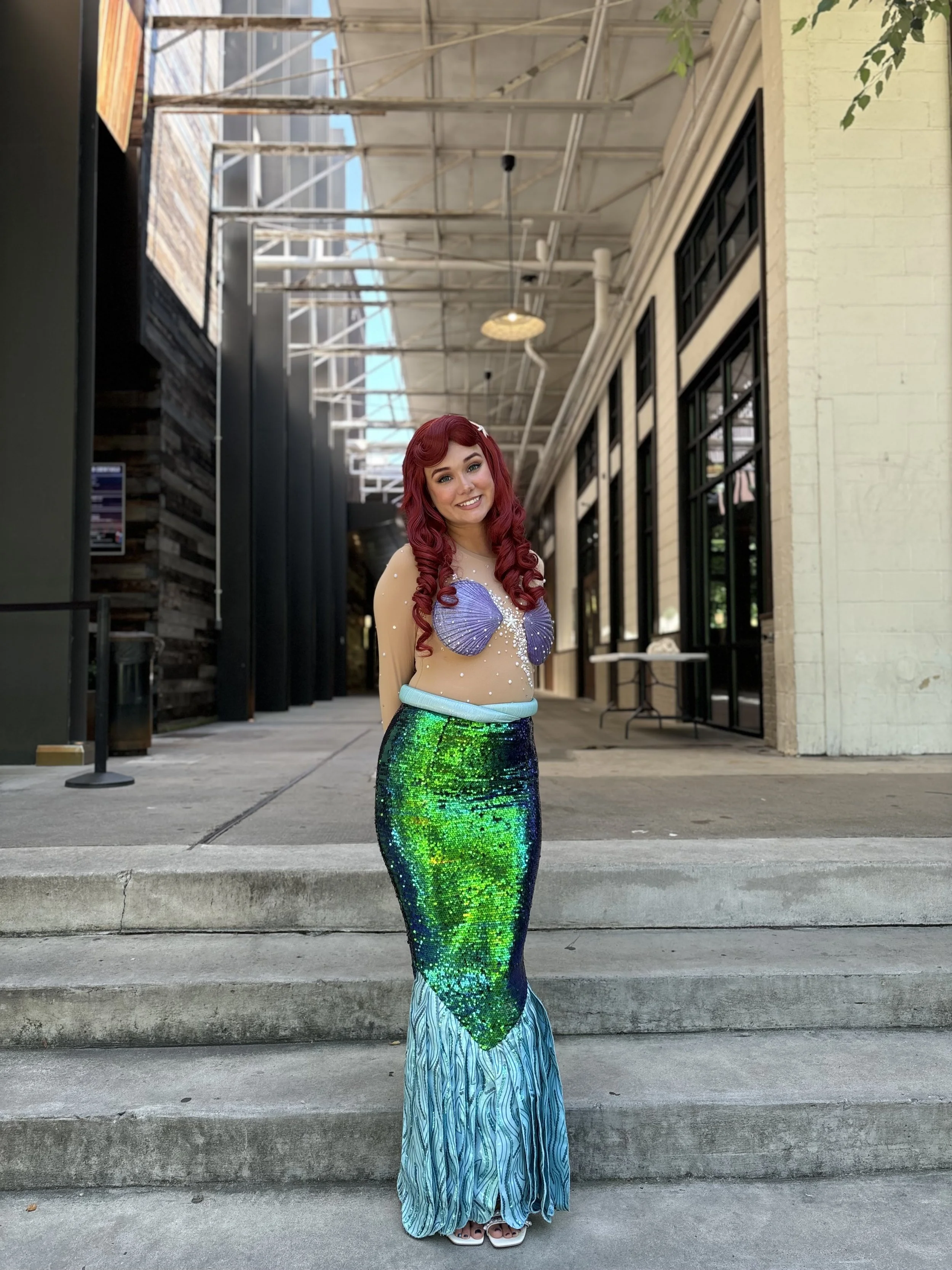 A woman standing on steps outside a building, dressed as a mermaid with red curly hair, a seashell top, and a shimmering green and blue tail.