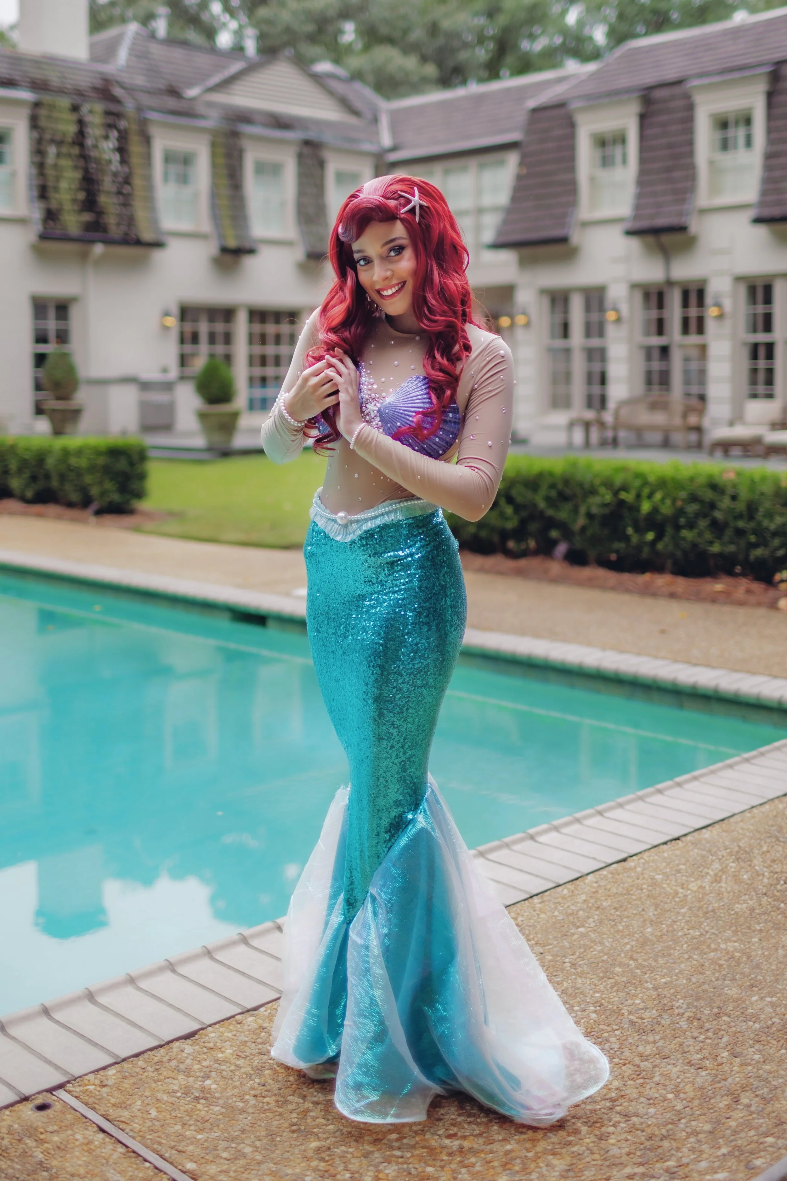 A woman dressed as Ariel, The Little Mermaid, standing by a swimming pool in a poolside area with residential buildings in the background.