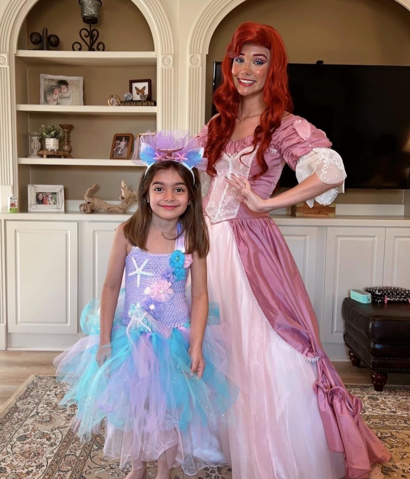 A woman dressed as a princess with long red hair and a young girl dressed as a mermaid with a pastel purple and blue tutu, standing in a living room with a television and decorative shelves in the background.