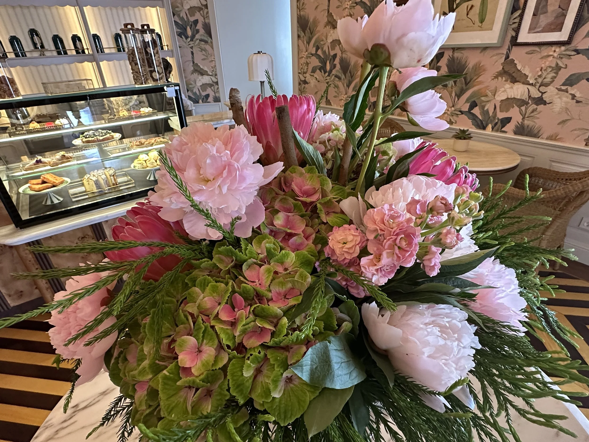 Close-up of a large, colorful flower bouquet with pink peonies, hydrangeas, and greenery, in a cozy cafe or bakery setting with a glass display case of desserts in the background.