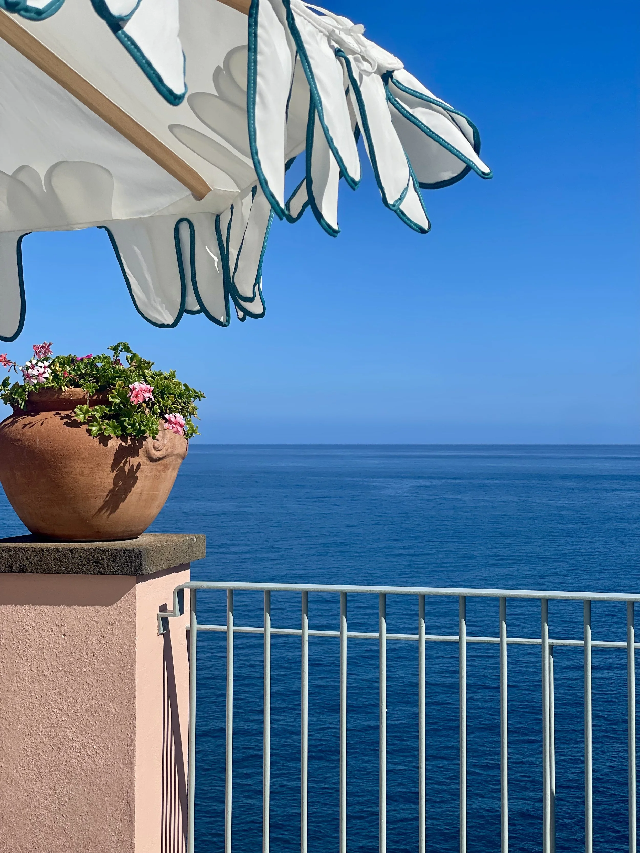 A terrace overlooking a calm, blue ocean, with a pink wall, a potted pink flower plant, a white and blue striped umbrella, and a metal railing.
