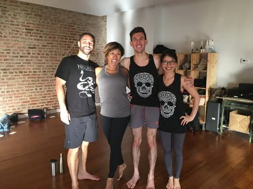 Four people standing together in a room, smiling for a group photo. The room has a brick wall and wooden flooring, with shelves and various items in the background.