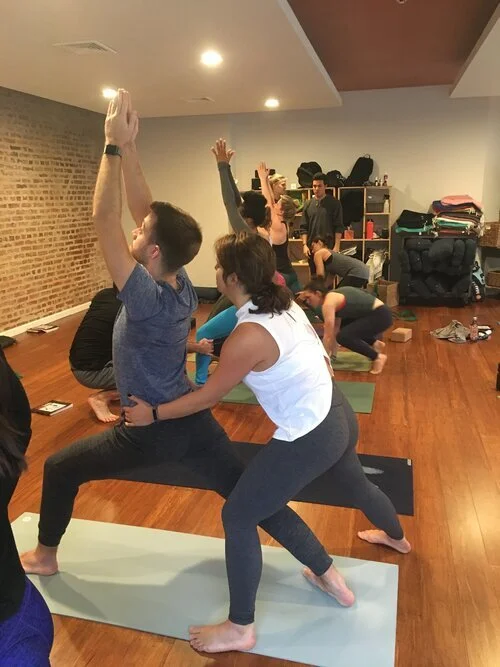 A group of people practicing yoga in a studio, with some in Warrior Pose and others in different poses, on mats with shelves and a brick wall in the background.