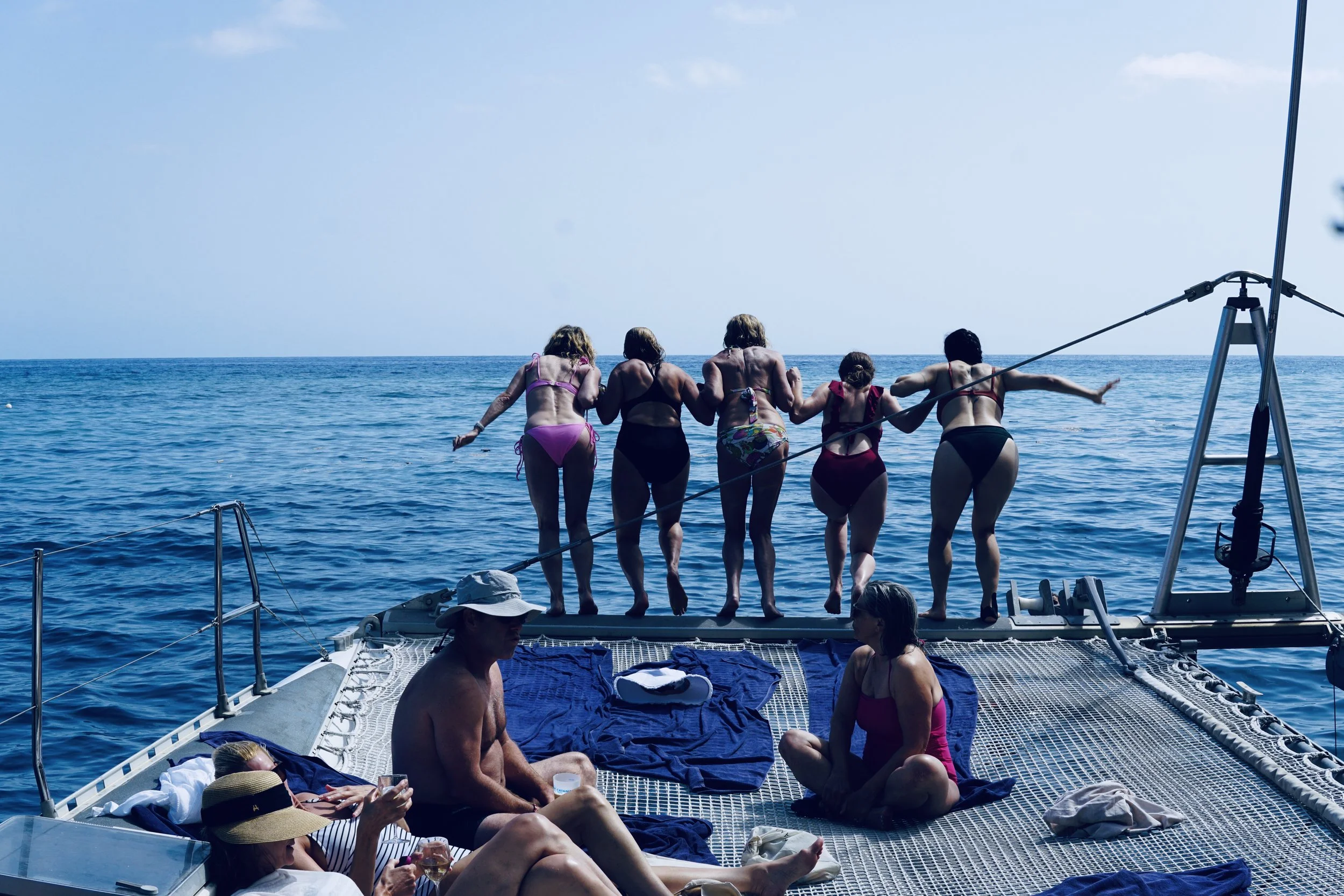 People relaxing on a boat deck and women standing on the edge holding a rope and enjoying the ocean view.
