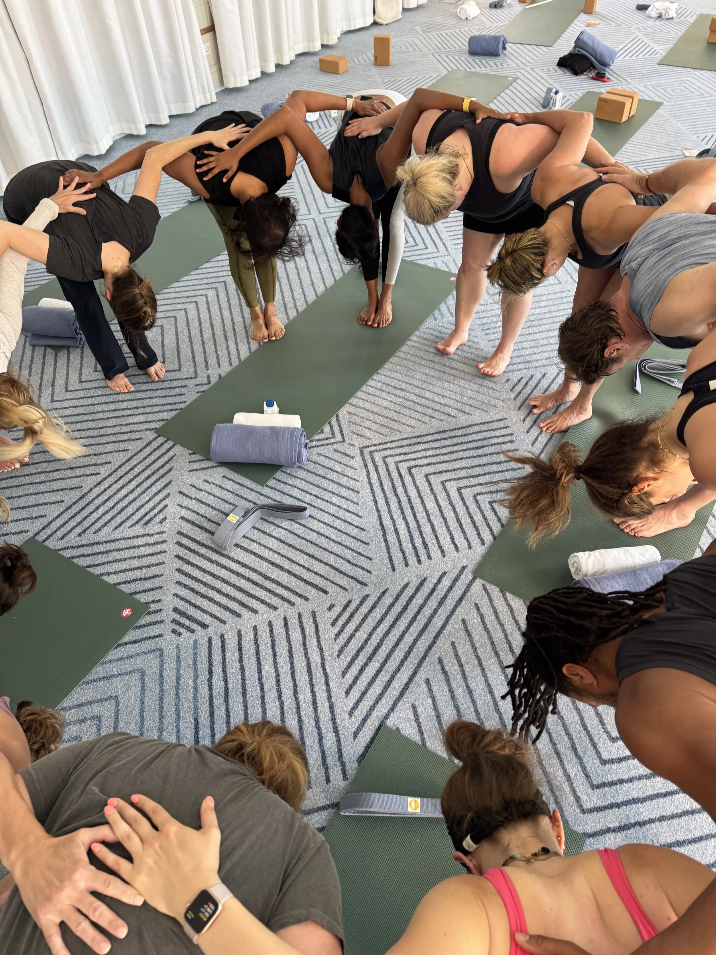 Group of people practicing yoga in a circle on mats in a room with geometric carpet pattern, yoga blocks, and towels.
