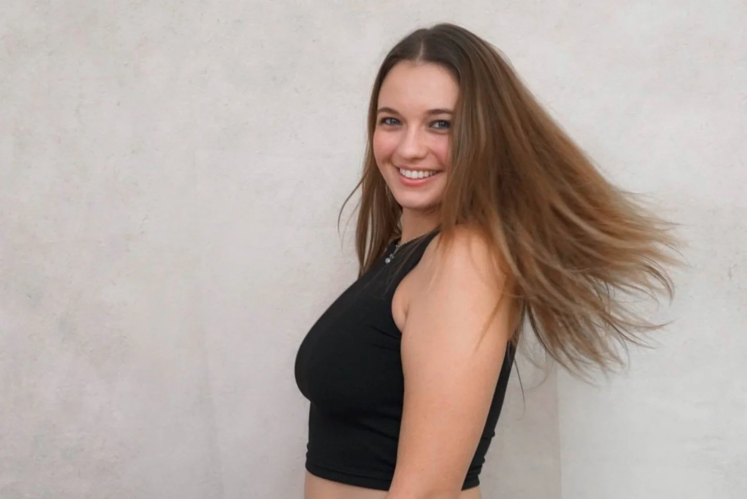 A woman with long, flowing brown hair and a smile, turning to the side and wearing a black sleeveless top against a plain, light-colored background.
