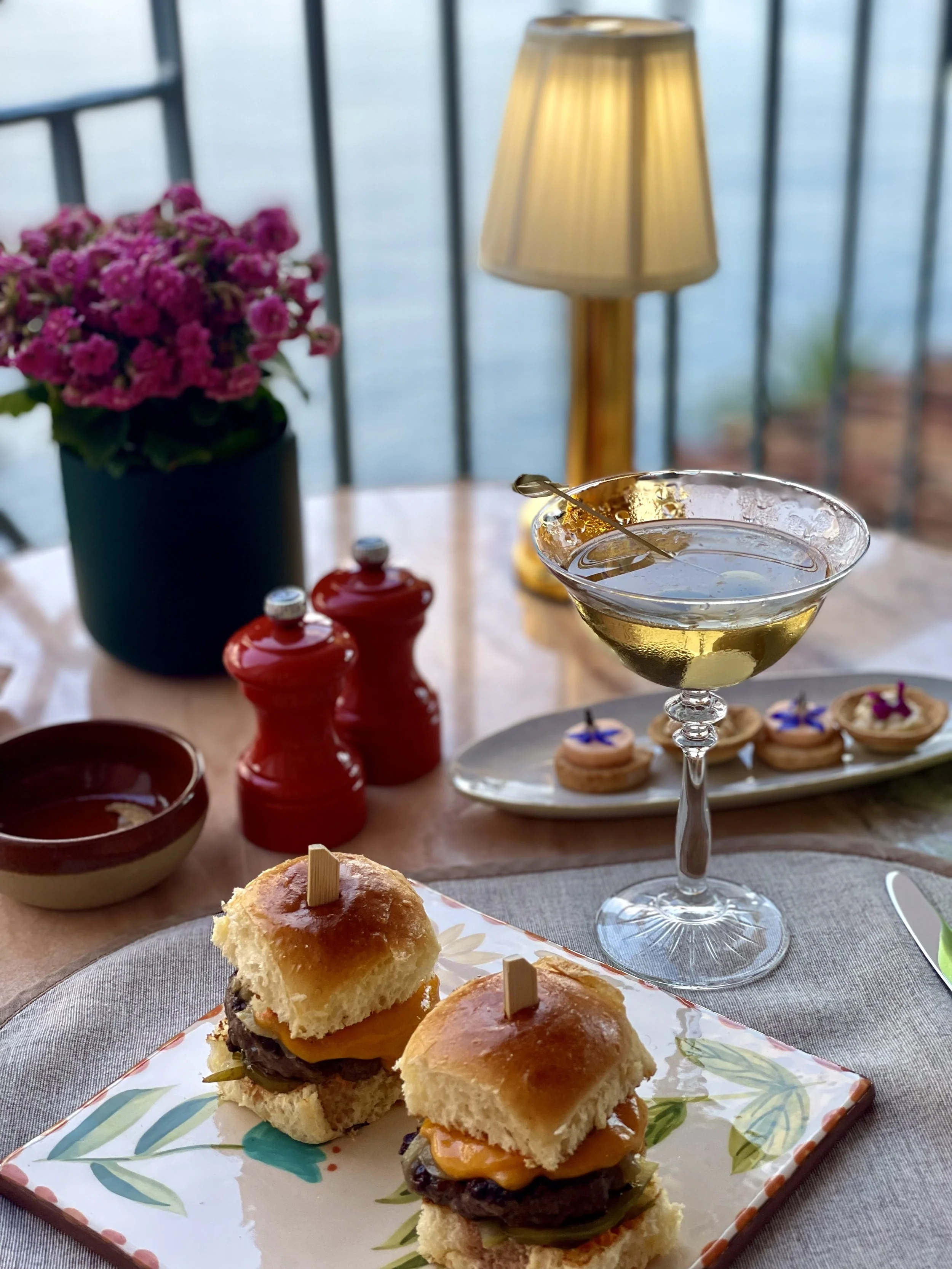 Table with two sliders, a cocktail glass with a lemon slice and a cocktail pick, a flowerpot with pink flowers, a lamp, salt and pepper shakers, and small desserts on a plate.