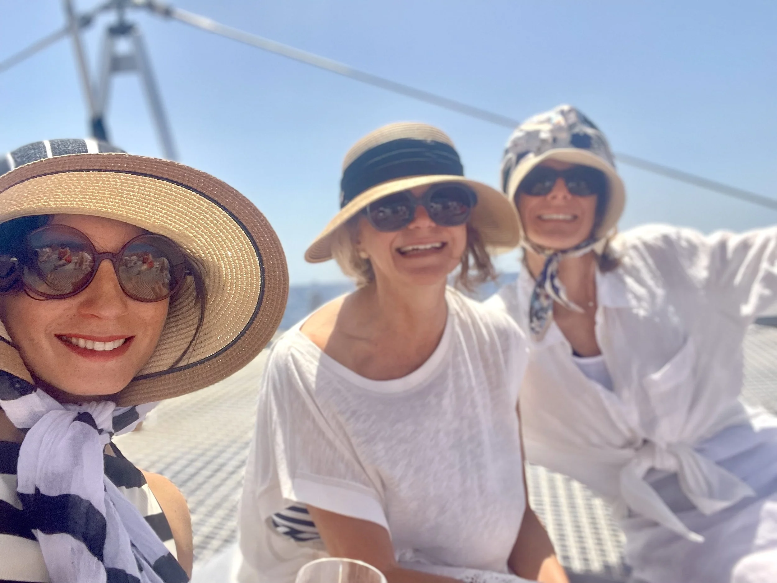 Three women wearing sun hats and sunglasses, smiling and enjoying a boat ride on a sunny day.