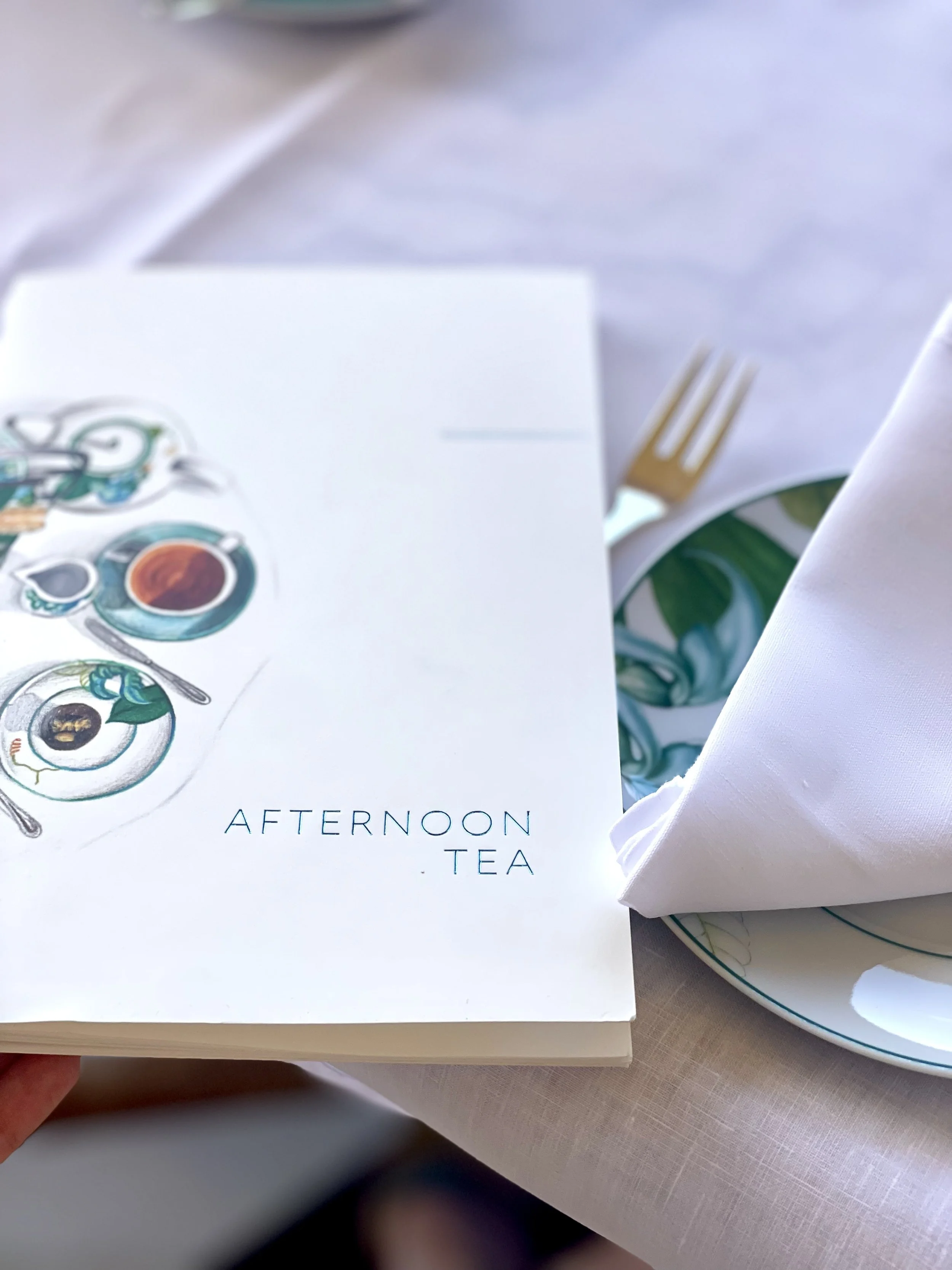 A menu titled 'Afternoon Tea' resting on a table with a white tablecloth, a plate with a green and white patterned napkin, a gold fork, and a white cloth napkin.