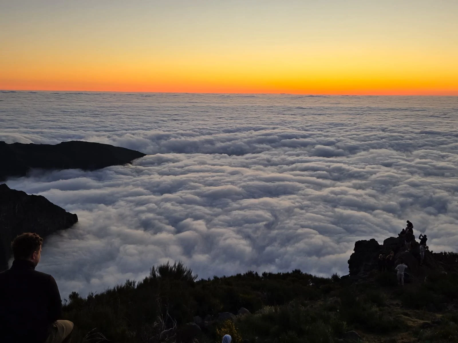 People sitting on rocky mountain ledge above the clouds during sunset with orange and yellow sky, dark silhouette of mountains and trees.