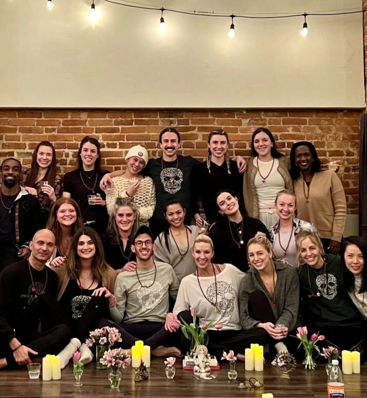 A group of 17 people gathered together smiling for a photo indoors against a brick wall with string lights hanging overhead. The group includes men and women of diverse backgrounds. In front of them is a table with flowers, candles, and small decorative items.