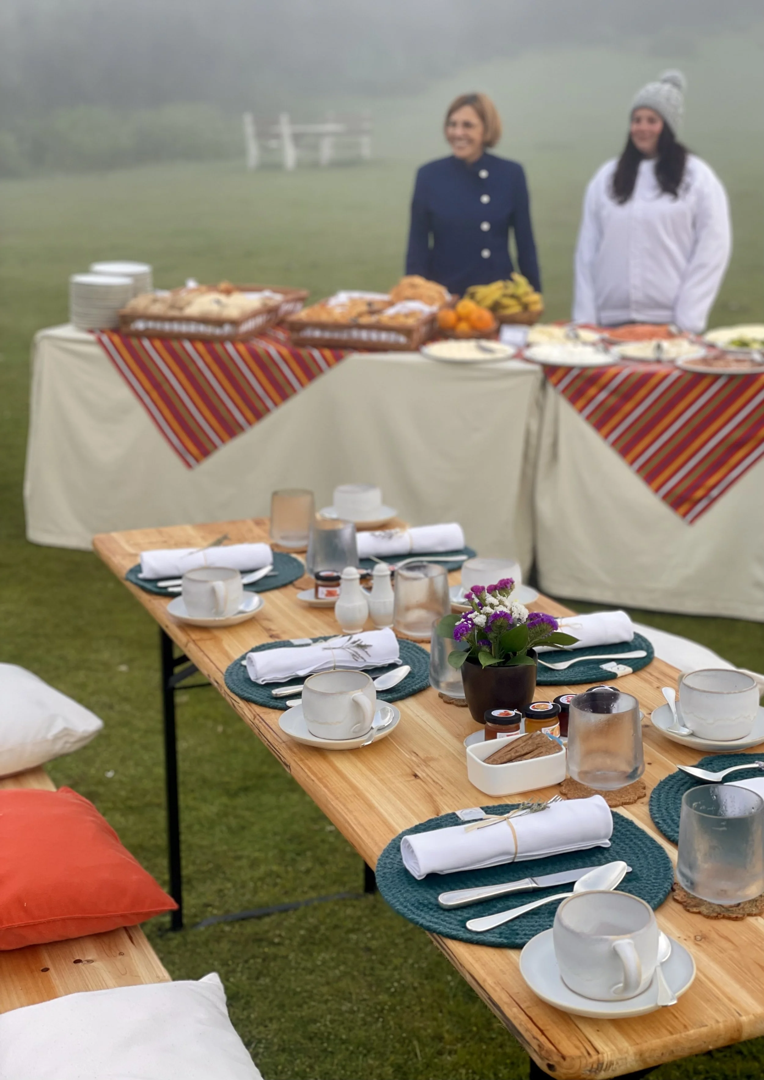 Outdoor breakfast setup with a wooden table, cups, glasses, napkins, and cutlery, next to a pillow on a grassy field, with a buffet table of food and two women standing behind it, one wearing a blue jacket and the other in a white jacket and gray kni