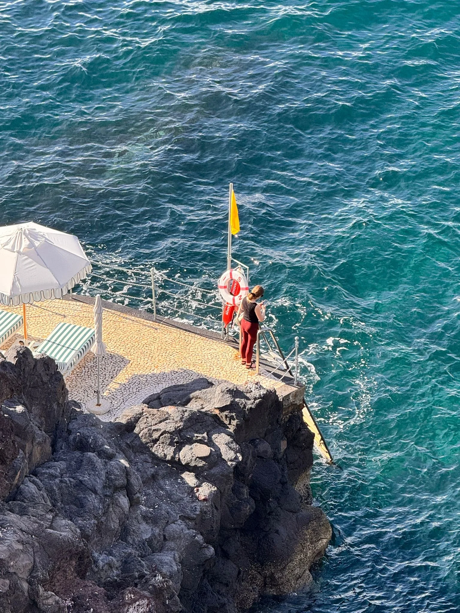 A woman standing at the edge of a concrete and rocky dock area, looking at the water, with an umbrella and lounge chairs nearby.