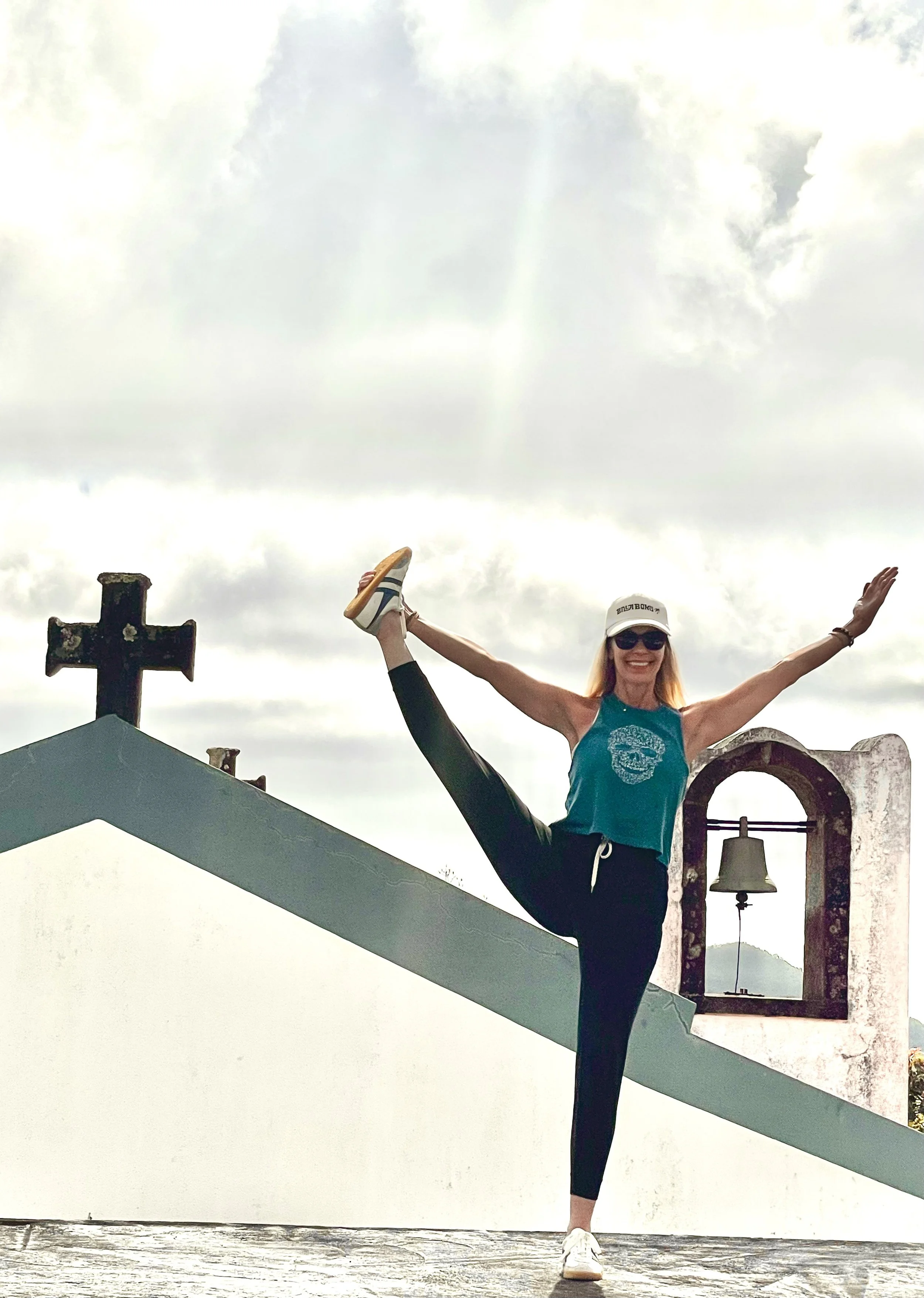 A woman performing a yoga pose on a rooftop, balancing on one foot while holding the other foot with her hand. She is smiling, wearing sunglasses, a white cap, a blue sleeveless top, and black leggings. The background shows an overcast sky and a chur