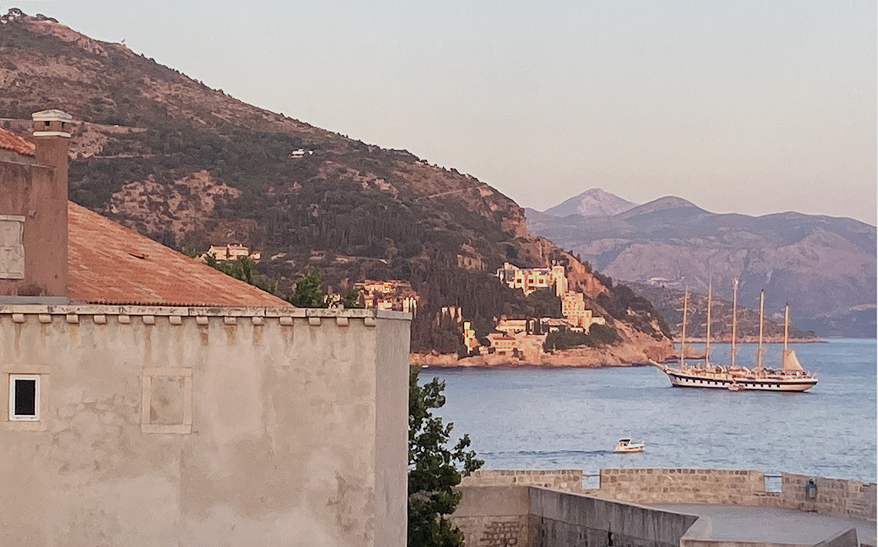A view of a coastal town with a sailboat on the water, mountains in the background, and buildings along the shoreline at sunset.