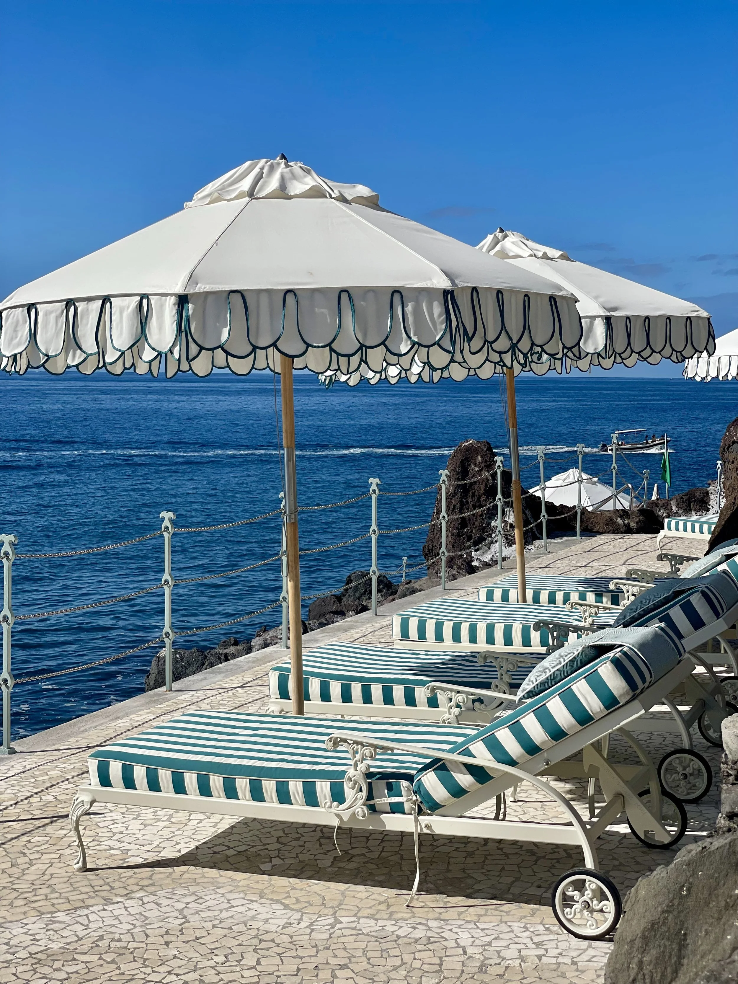 Beach chairs with blue and white striped cushions and open white umbrellas on a coastal promenade with a view of the sea and boats.