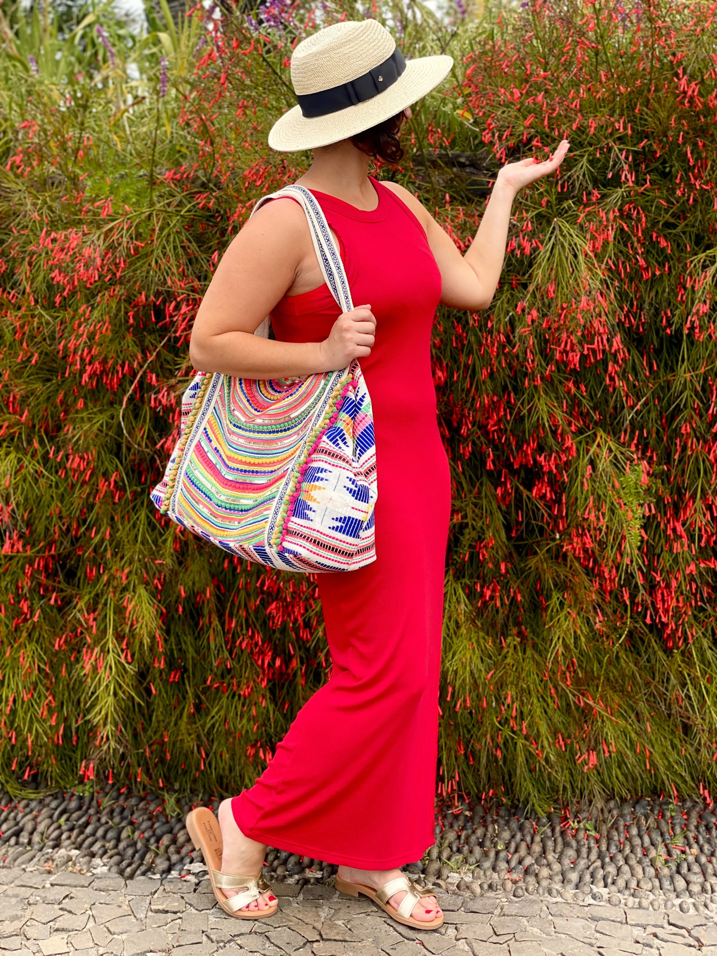 Woman in a red maxi dress and straw hat standing in front of red flowering bushes, carrying a colorful patterned bag.