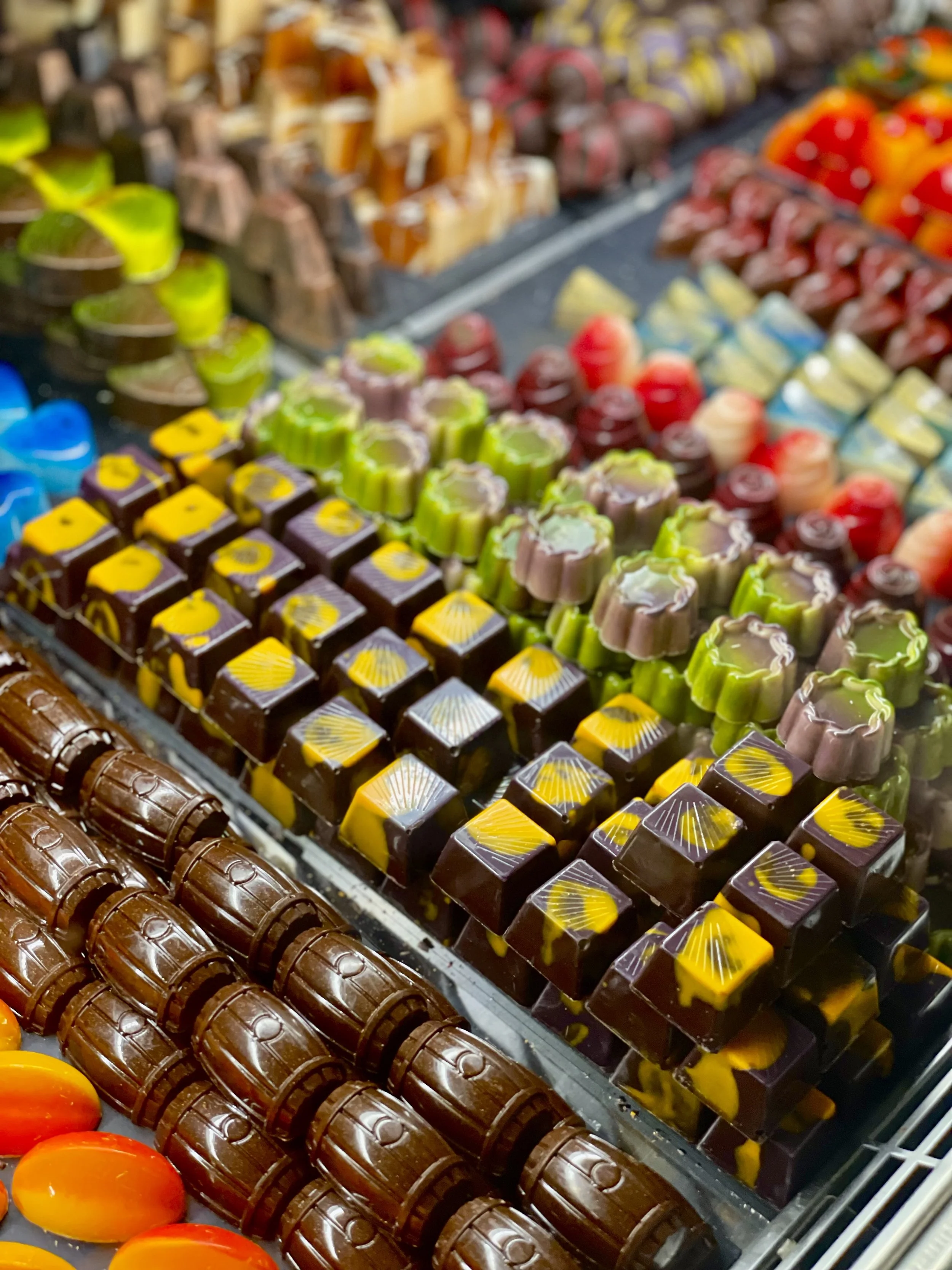 A display of various colorful chocolates arranged in rows at a store or market. The chocolates are in different shapes and colors, including green, yellow, brown, red, and purple.