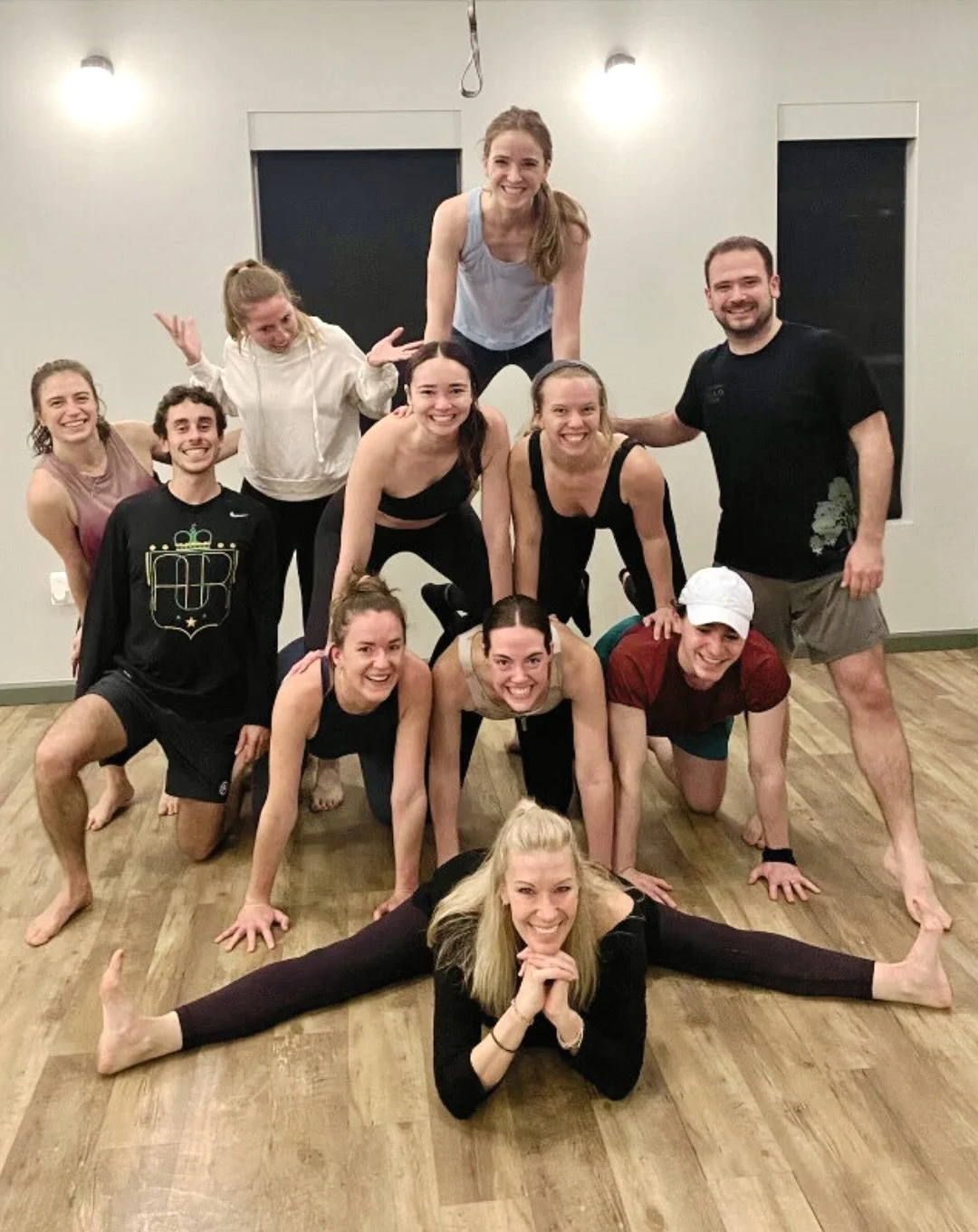 A group of eleven people, posing in a gym or dance studio, forming a human pyramid and performing a split on the floor, all smiling and looking happy.
