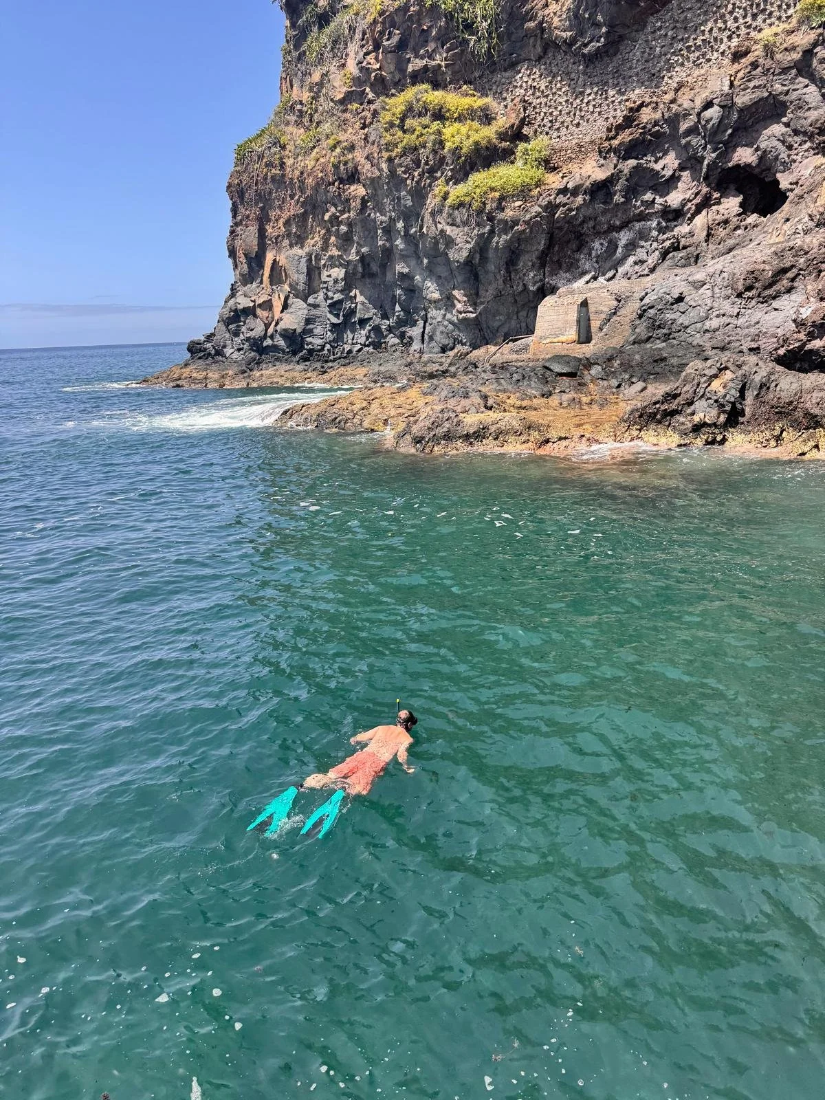 A person snorkeling in clear waters near a rocky coastline with cliffs and sparse vegetation.