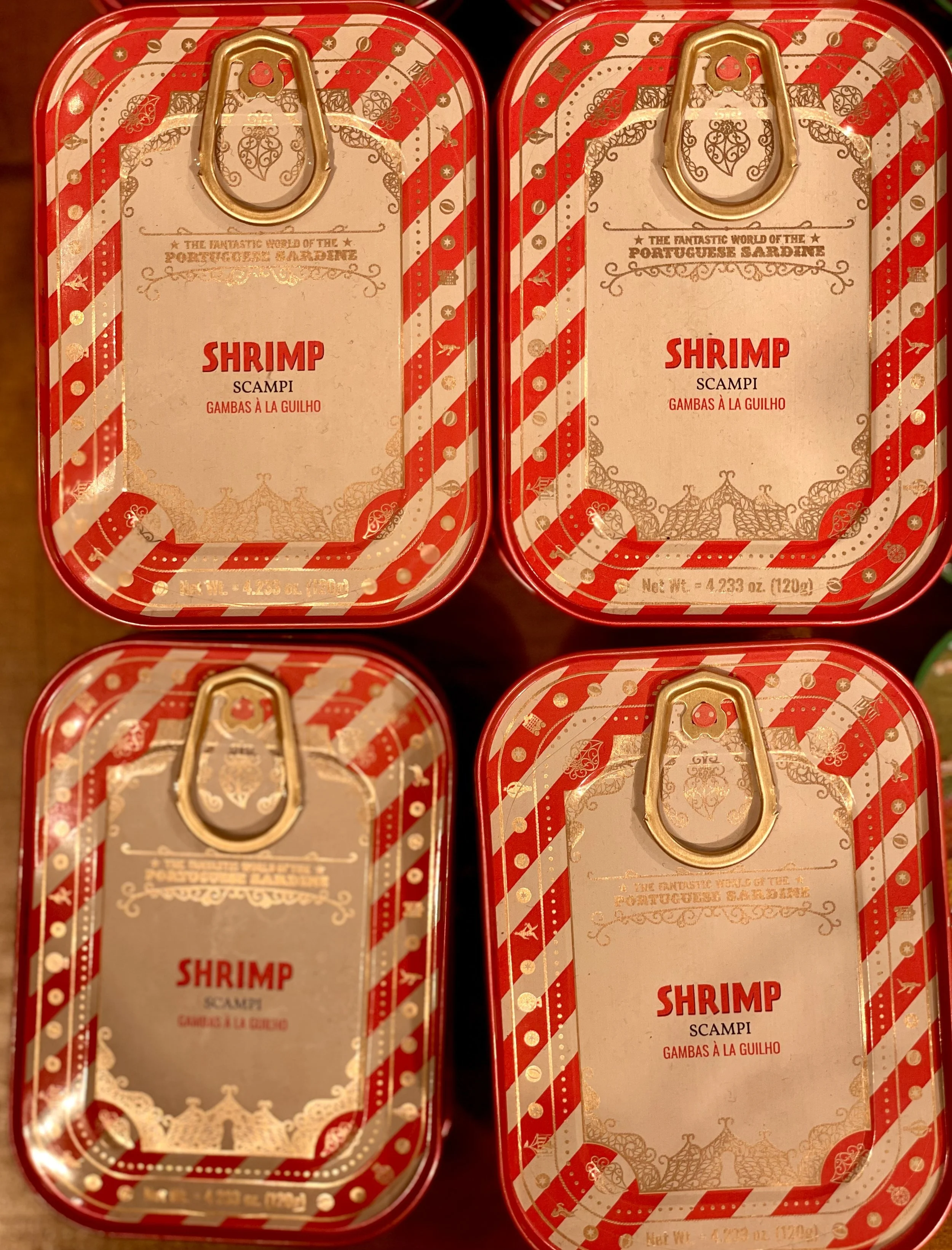 Four red and white striped tins of Portuguese sardines with gold pull tabs, labeled 'Shrimp Scampi' in red text, with decorative patterns and text in English and Portuguese.