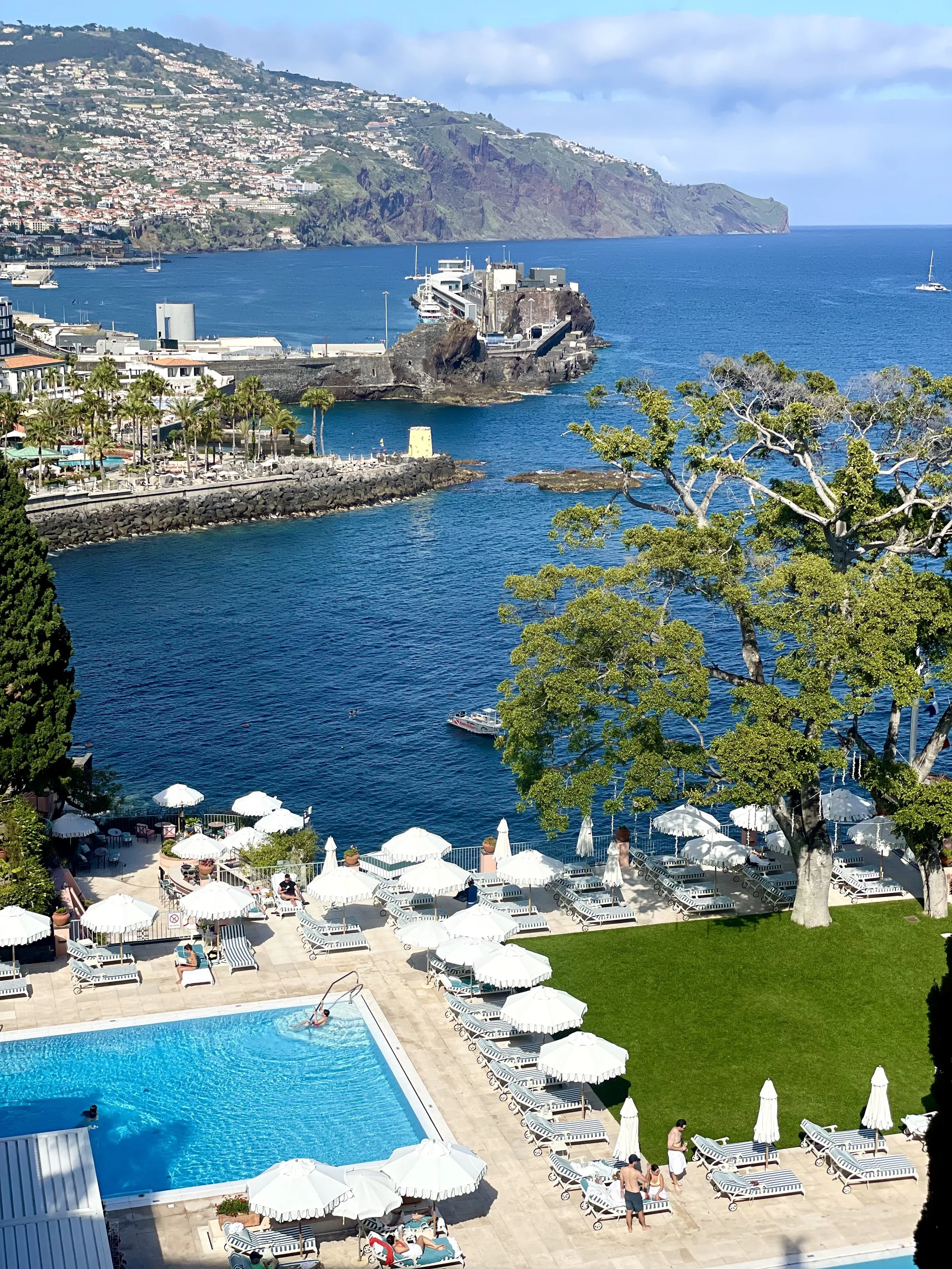 Pool area with white umbrellas and lounge chairs overlooking a harbor with boats, rocky coastline, and hillside buildings in the background.