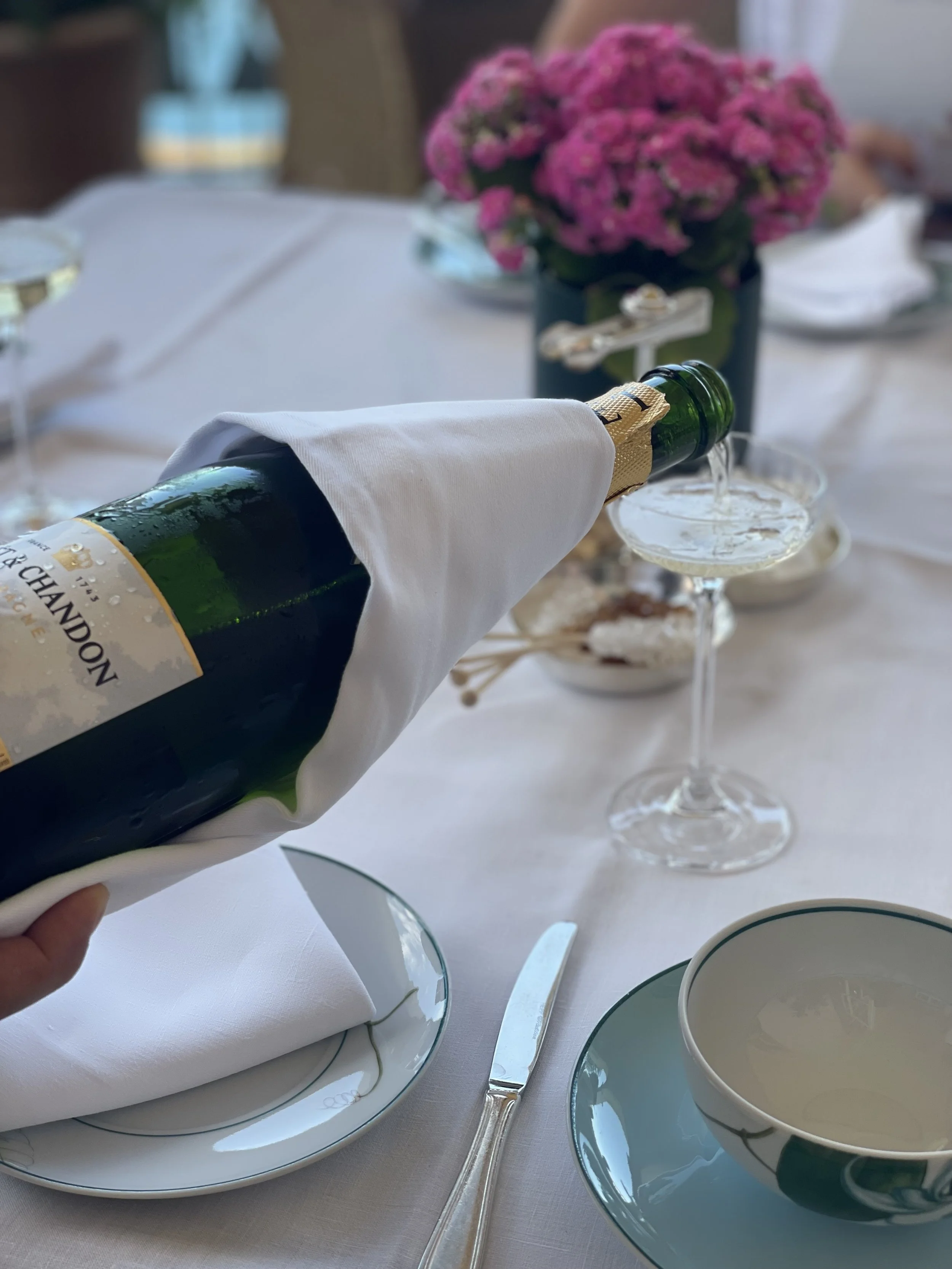 A person pouring champagne into a glass at a formal dining table decorated with a pink flower arrangement.