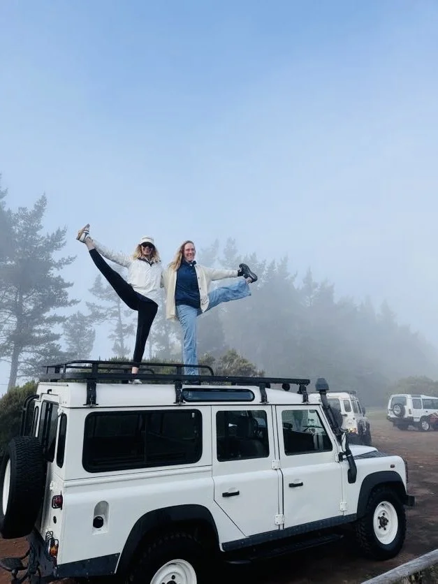 Two women on top of a white off-road vehicle, wearing athletic clothes and doing yoga poses with a forest and foggy sky in the background.