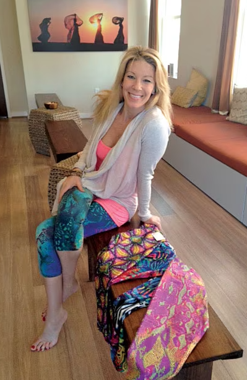 A woman with blonde hair, smiling, seated on a wooden bench in a cozy room with colorful fabrics spread out in front of her. The room has natural light coming through the windows, a framed art piece on the wall, and a window seat with pillows.