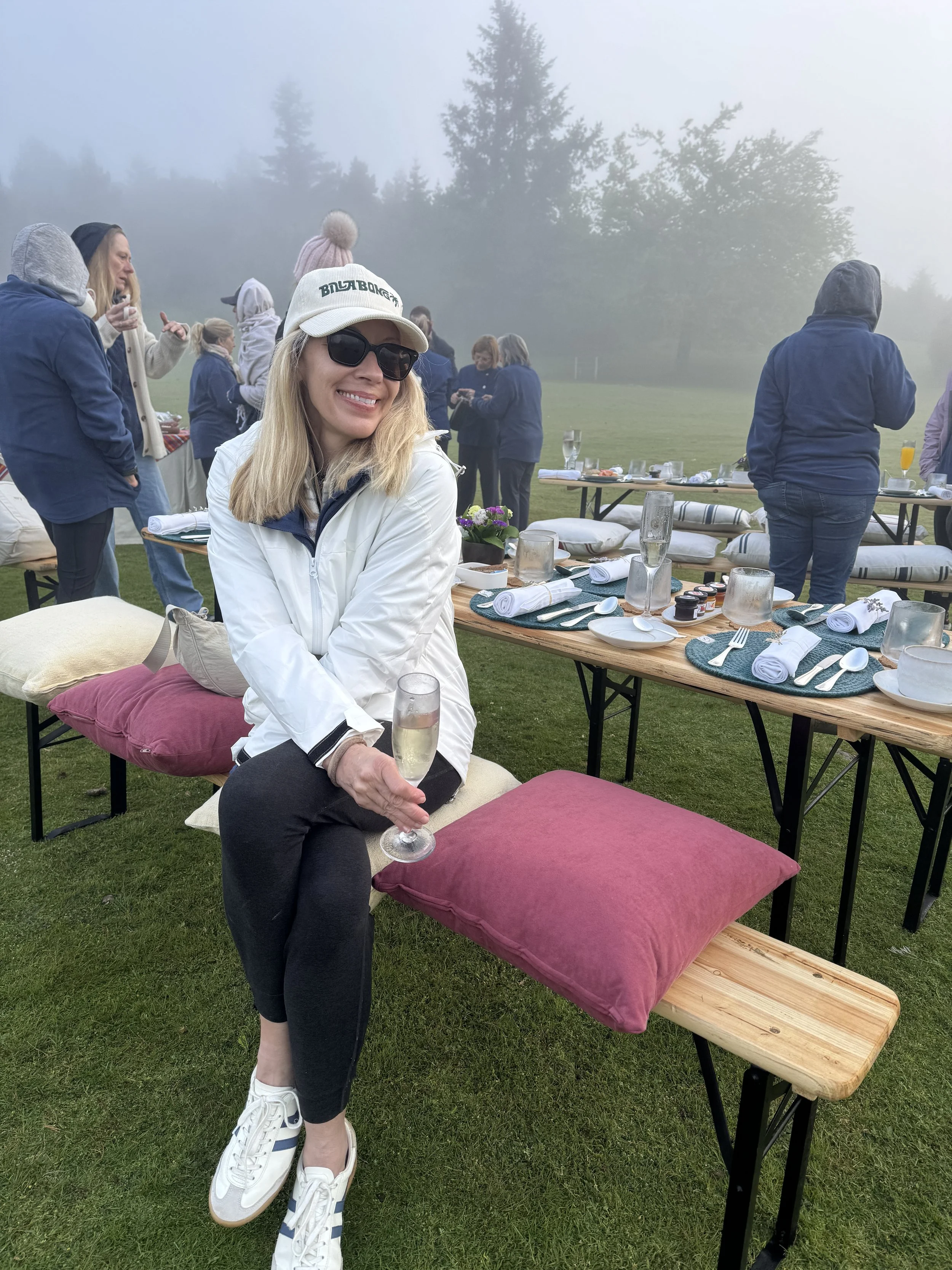 A woman sitting on a bench at an outdoor gathering, holding a glass of champagne, wearing sunglasses and a white jacket with a white cap, surrounded by a table set for a meal with multiple people in the background on a foggy day.