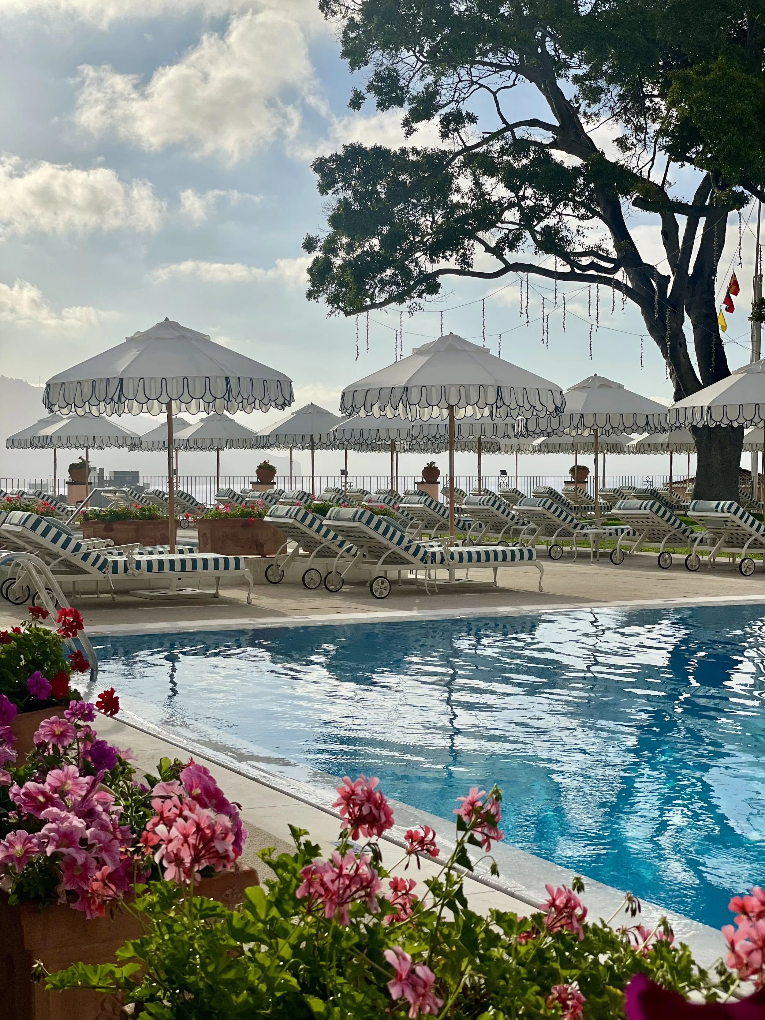 Poolside scene with striped lounge chairs and umbrellas, potted flowers, a large tree, and a partly cloudy sky.