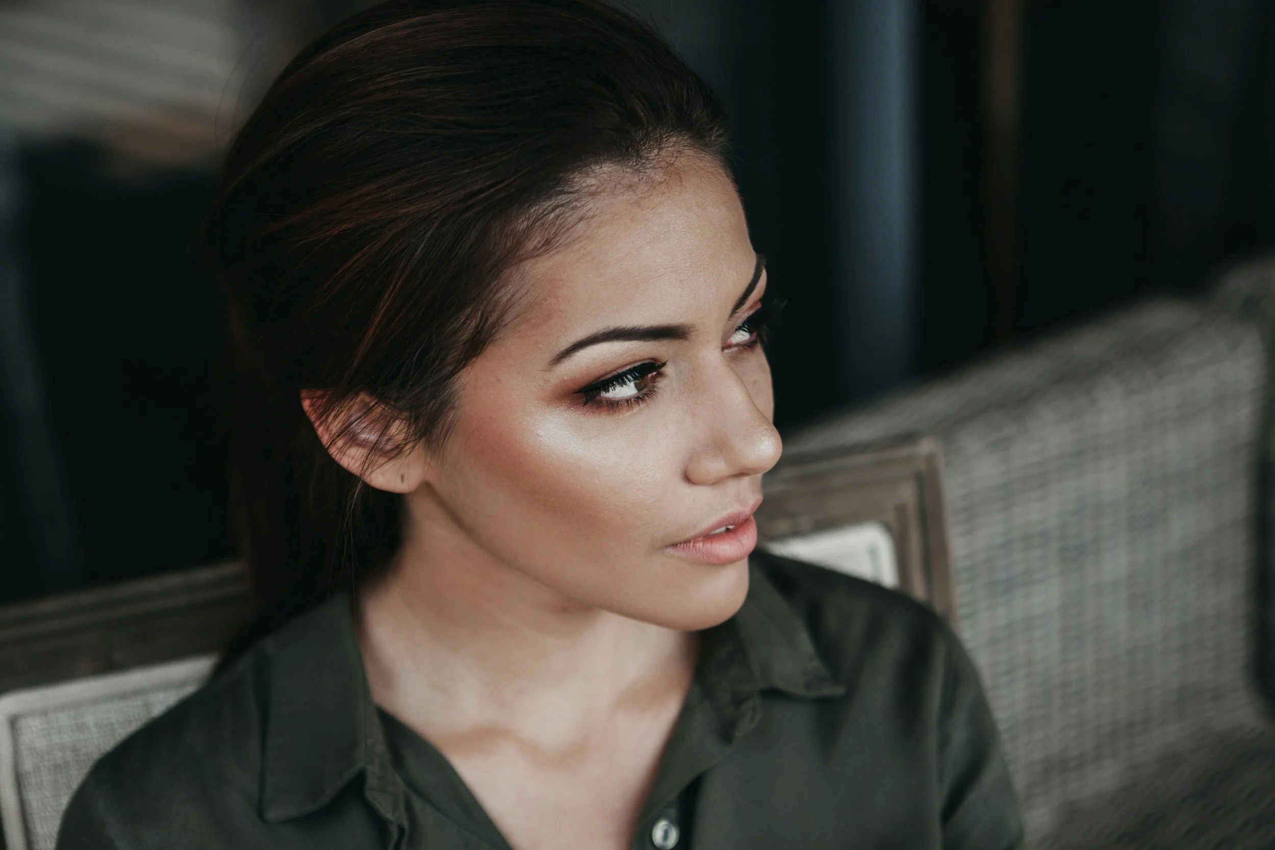 Close-up of a woman's face with makeup, sitting in an office or workspace, with her hair styled back and wearing a black shirt.