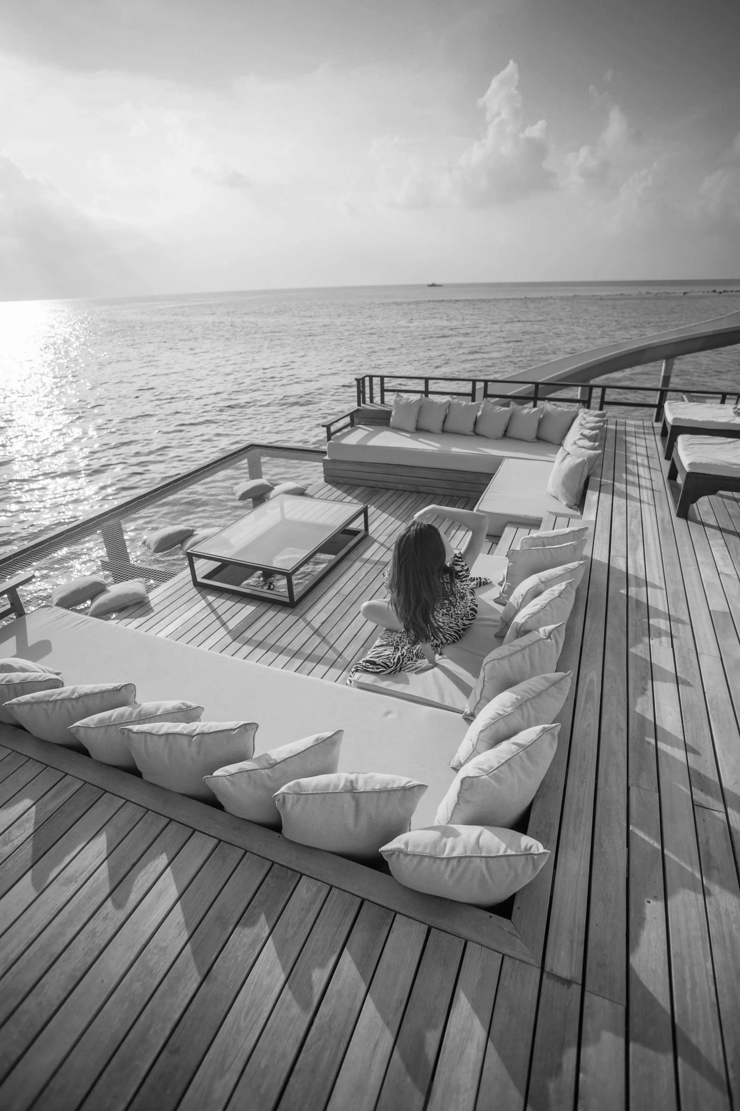 A woman sitting on an outdoor lounge area on a wooden deck by the water, with pillows, overlooking the ocean and cloudy sky.