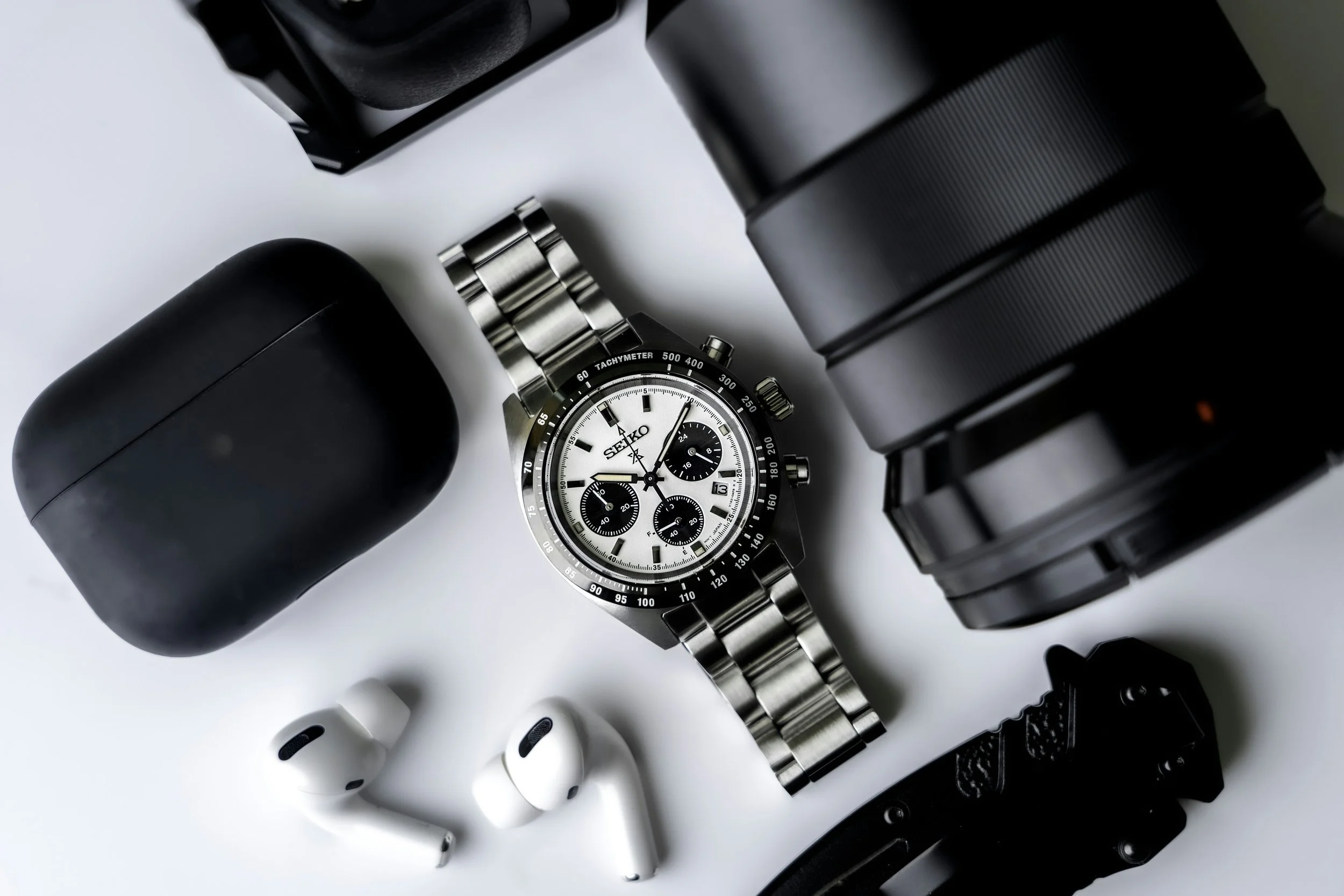 Close-up of a silver Seiko chronograph wristwatch with a white dial, surrounded by black camera lenses and white wireless earbuds on a white surface.