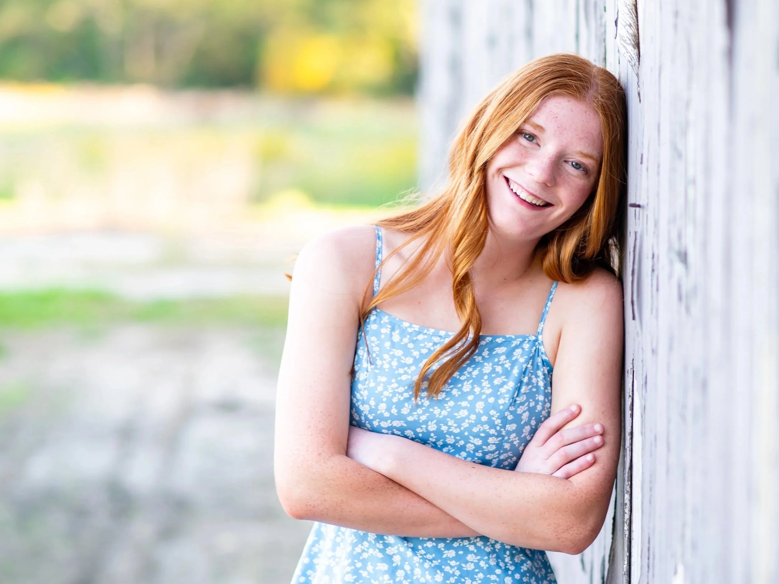 A young woman with red hair and freckles wearing a blue dress with white floral pattern, smiling and leaning against a white wooden wall outdoors on a sunny day.