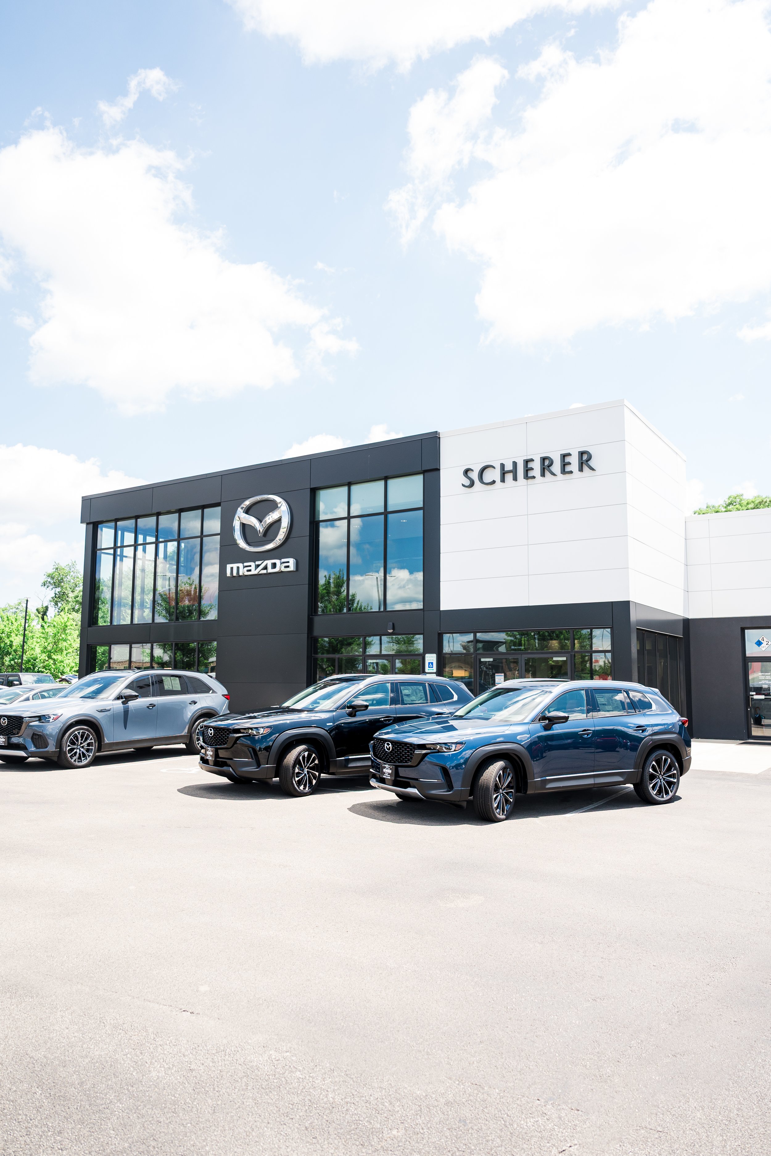 Mazda car dealership with multiple Mazda vehicles parked outside on a sunny day, modern building with Mazda logo and the name Scherer.