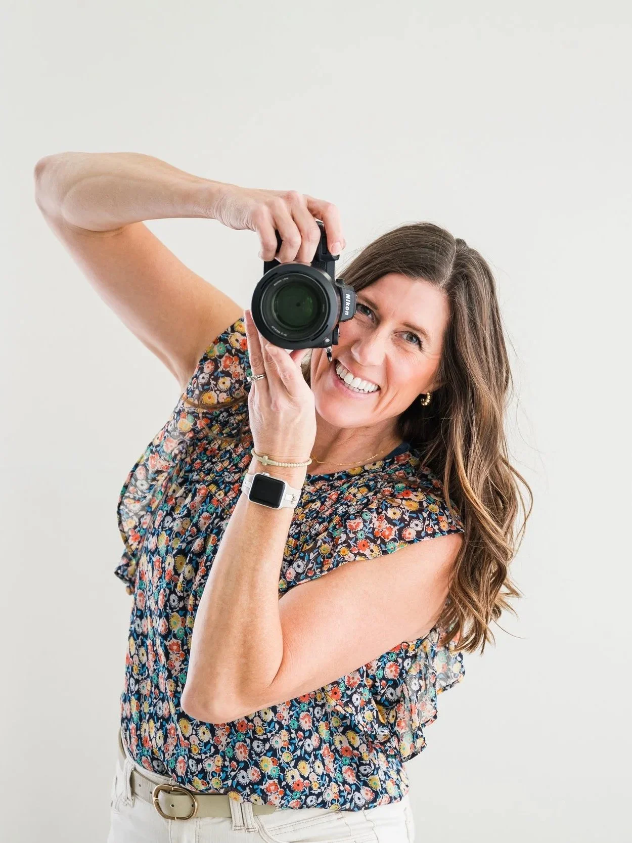A woman taking a selfie with a camera, smiling, wearing a floral top and wrist accessories, against a plain white background.