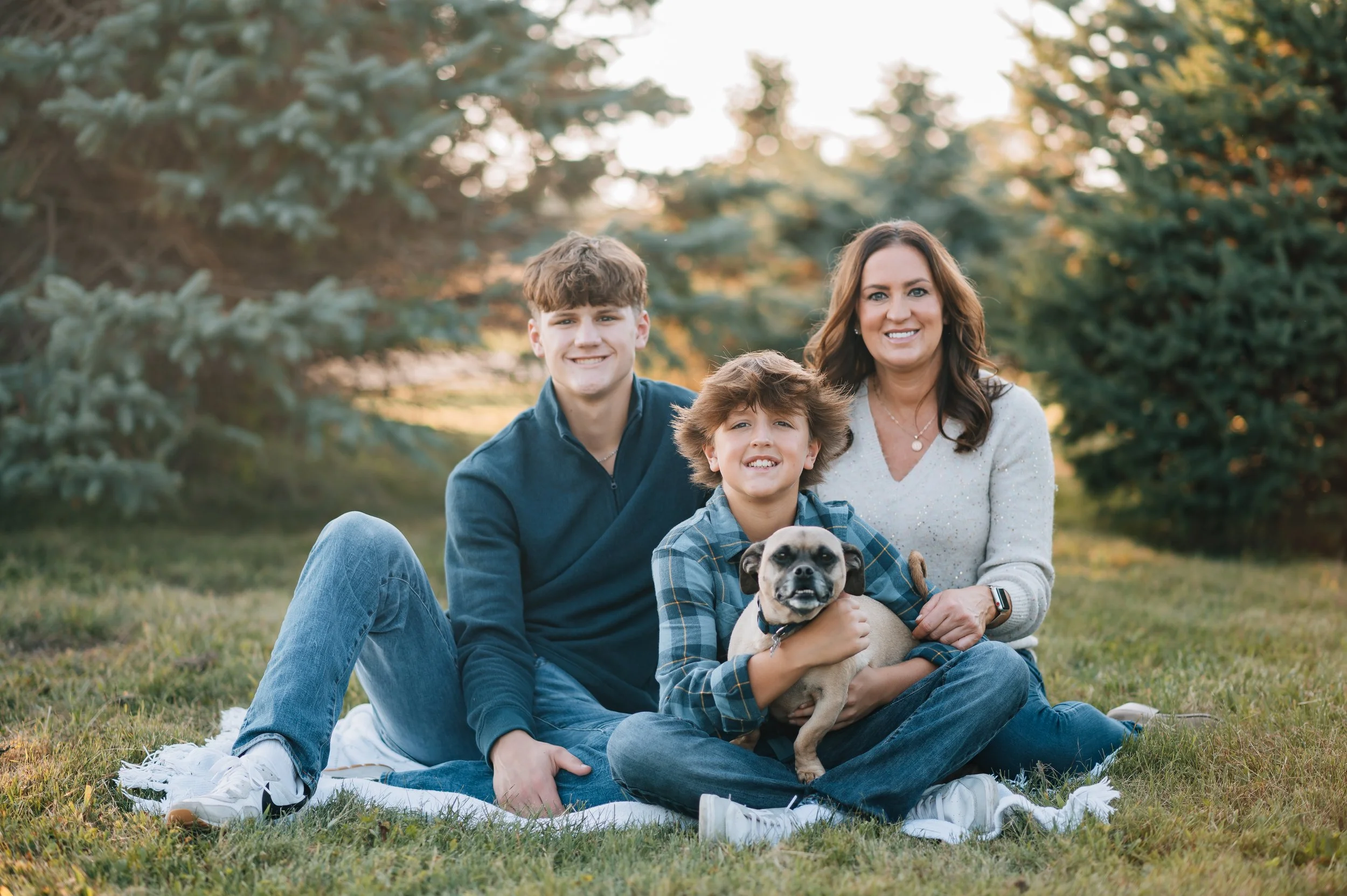 Family of three with a dog sitting on a blanket in a park with trees, smiling at the camera during sunset.