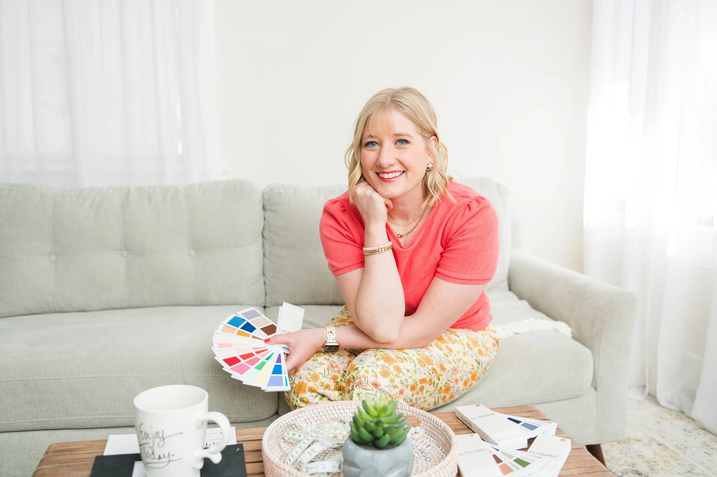 A woman with blonde hair and a pink shirt sitting on a light green sofa, smiling and holding color swatches, surrounded by samples and a coffee mug on a wooden table, with a succulent plant and sheer curtains in the background.