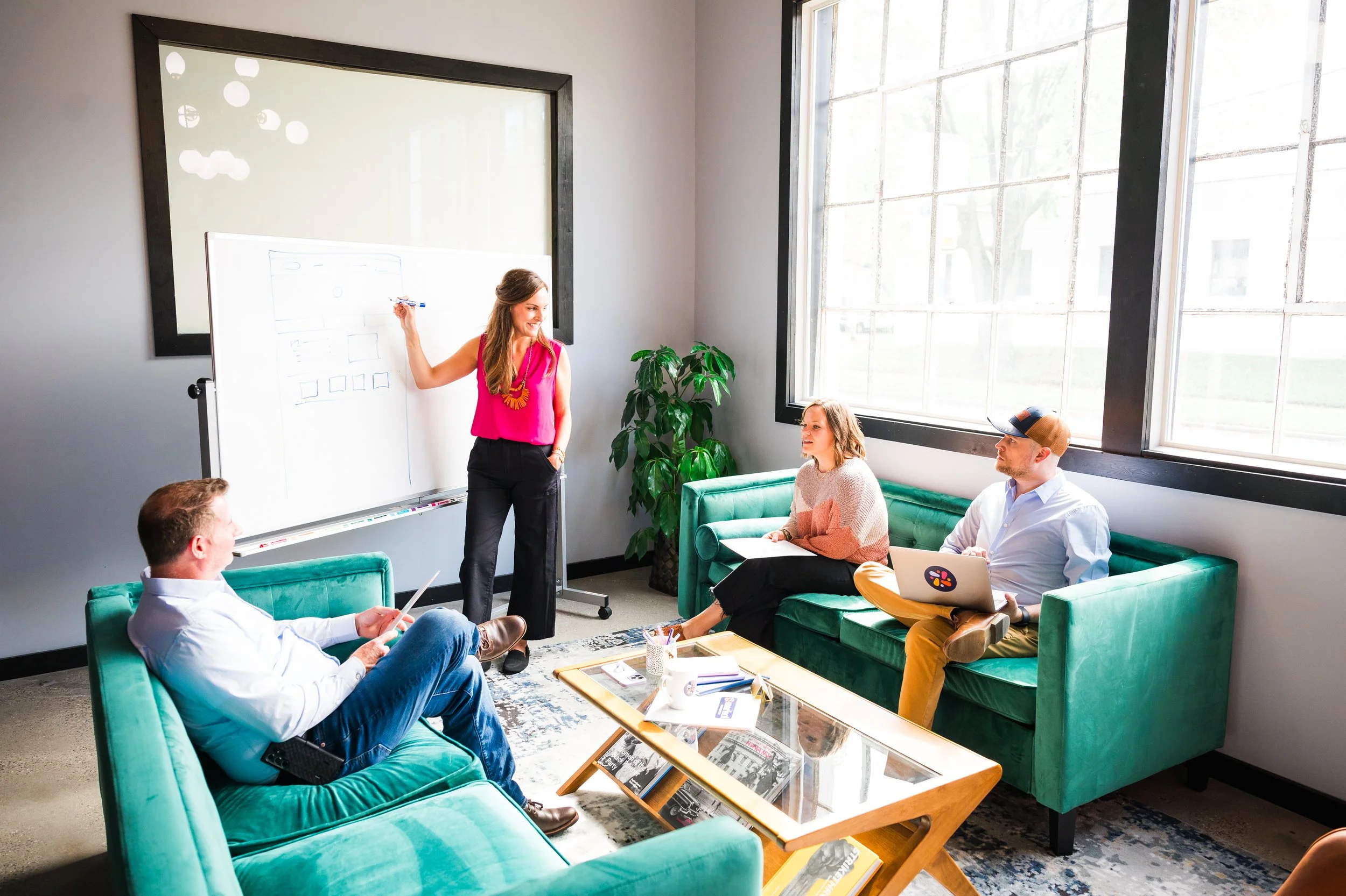 A woman presents to three colleagues in a modern, brightly lit office lounge with teal sofas, a whiteboard, and large windows, discussing a diagram.