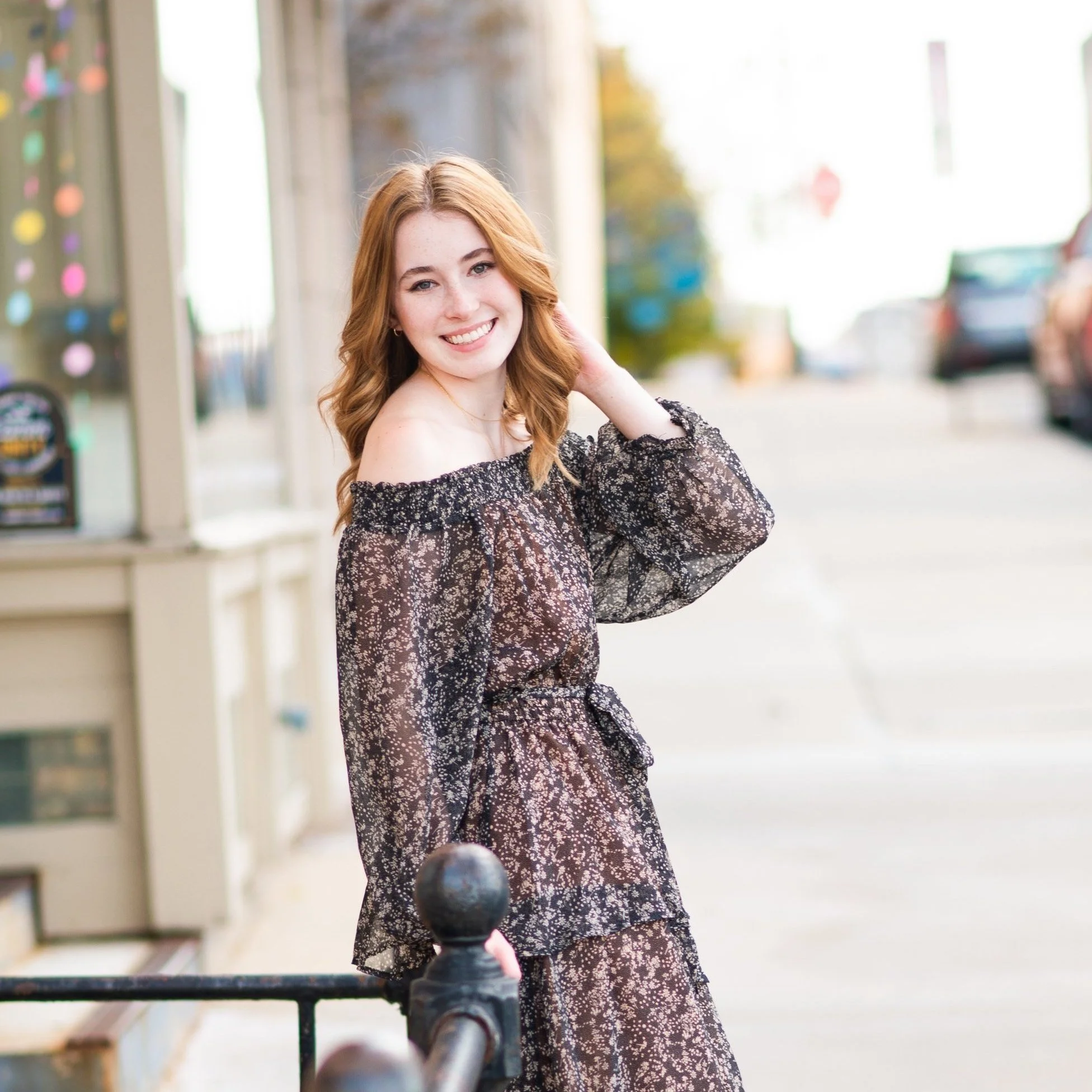 A young woman with reddish hair and a bright smile standing on a city sidewalk, wearing a black off-the-shoulder dress with white floral patterns.