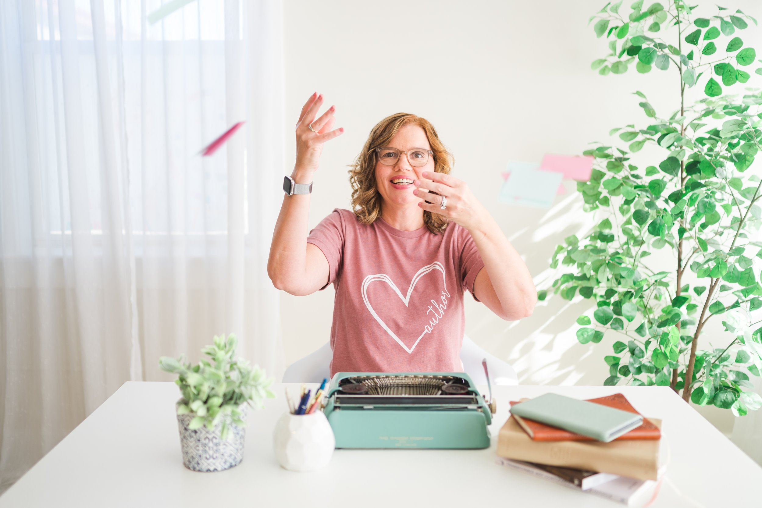 A woman with glasses, wearing a pink shirt with a heart and 'author' written inside, sitting at a white desk with a typewriter and various office supplies, gesturing animatedly with her hands.