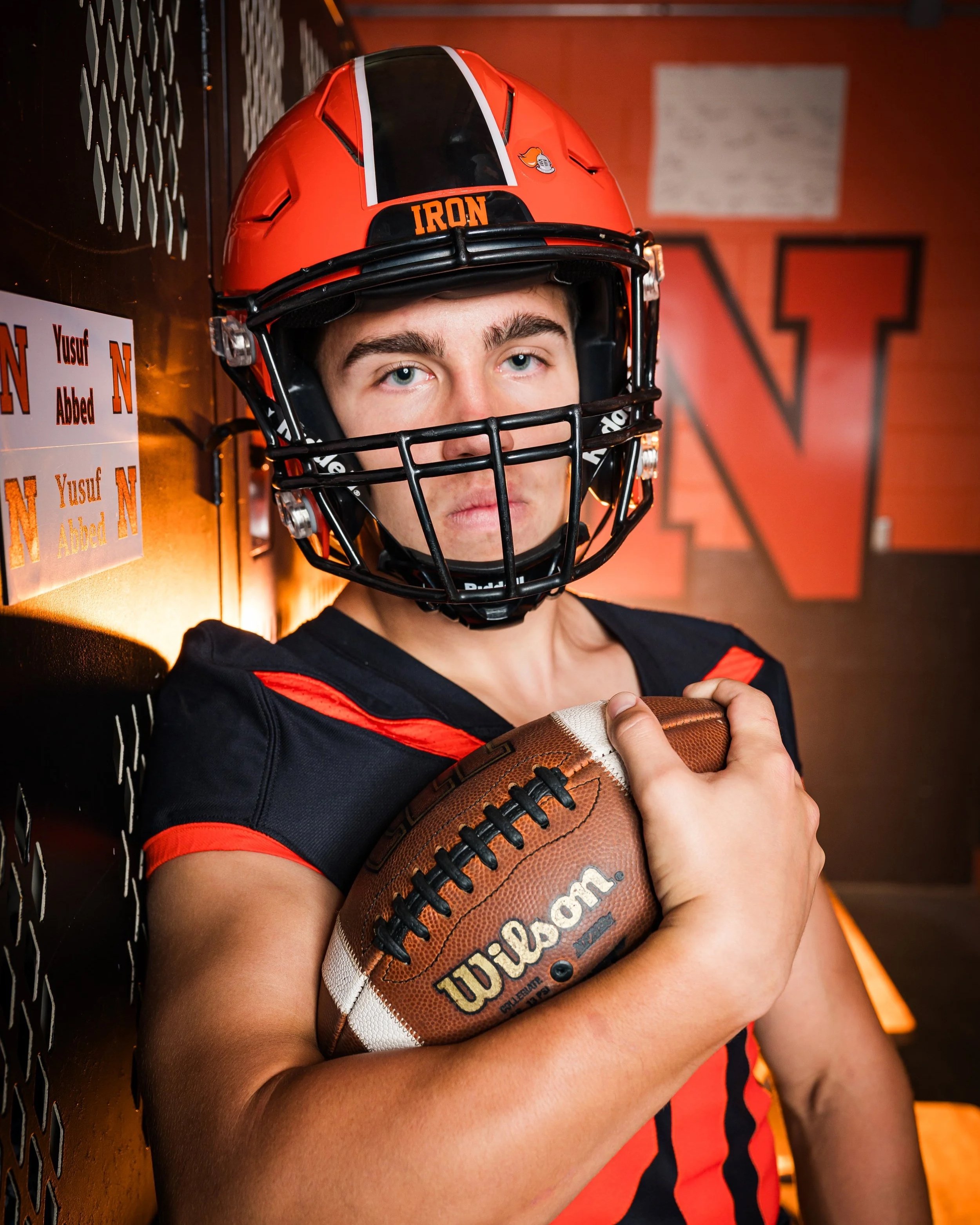 A young male football player wearing a black and orange helmet with a face guard, holding a Wilson football, standing in a locker room with a large letter 'N' on the wall and a name tag that reads 'Yusuf Abbed'.