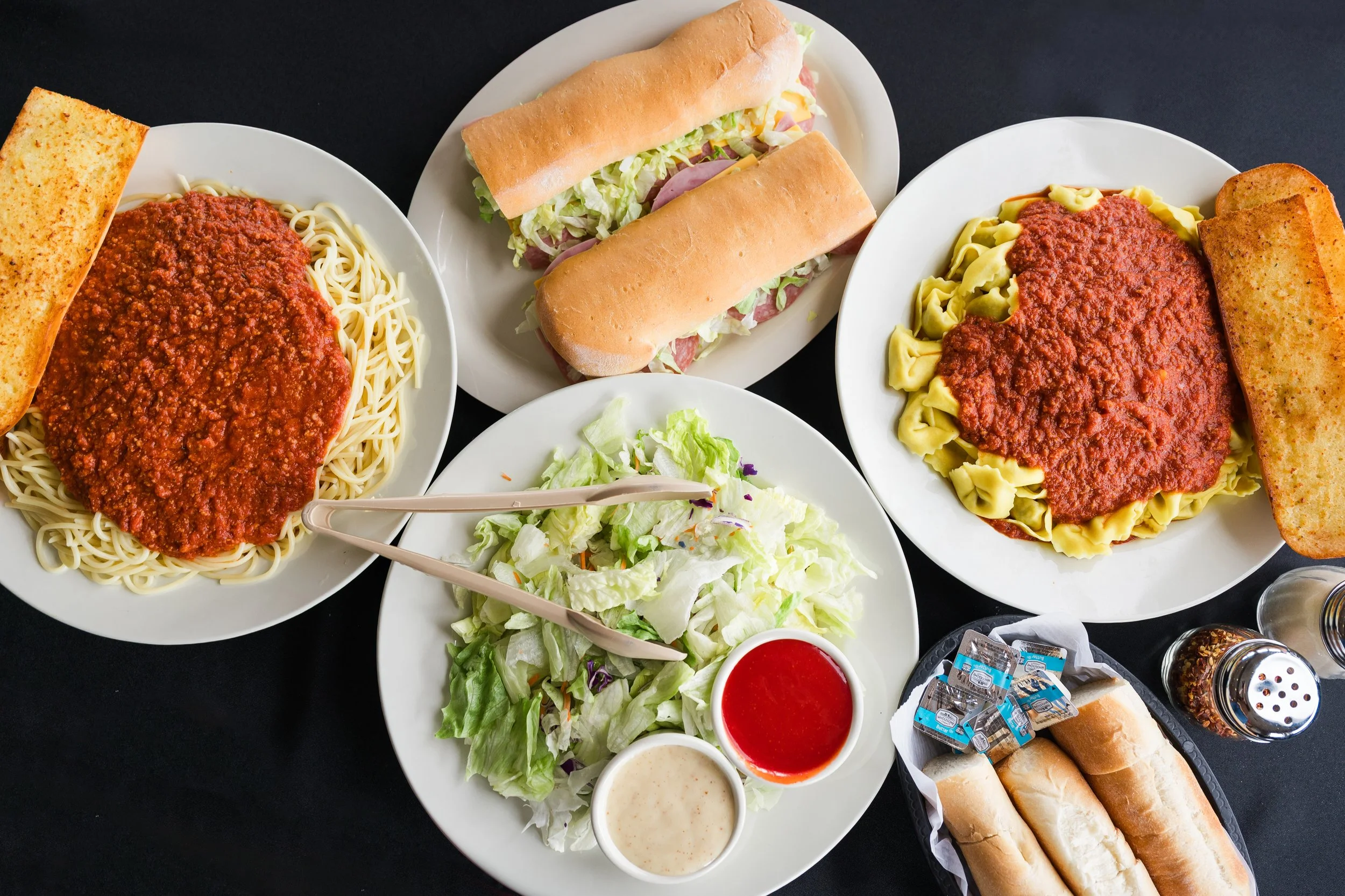 Pizza pasta, two submarine sandwiches, salad with dressings, garlic bread, and breadsticks on a black table.