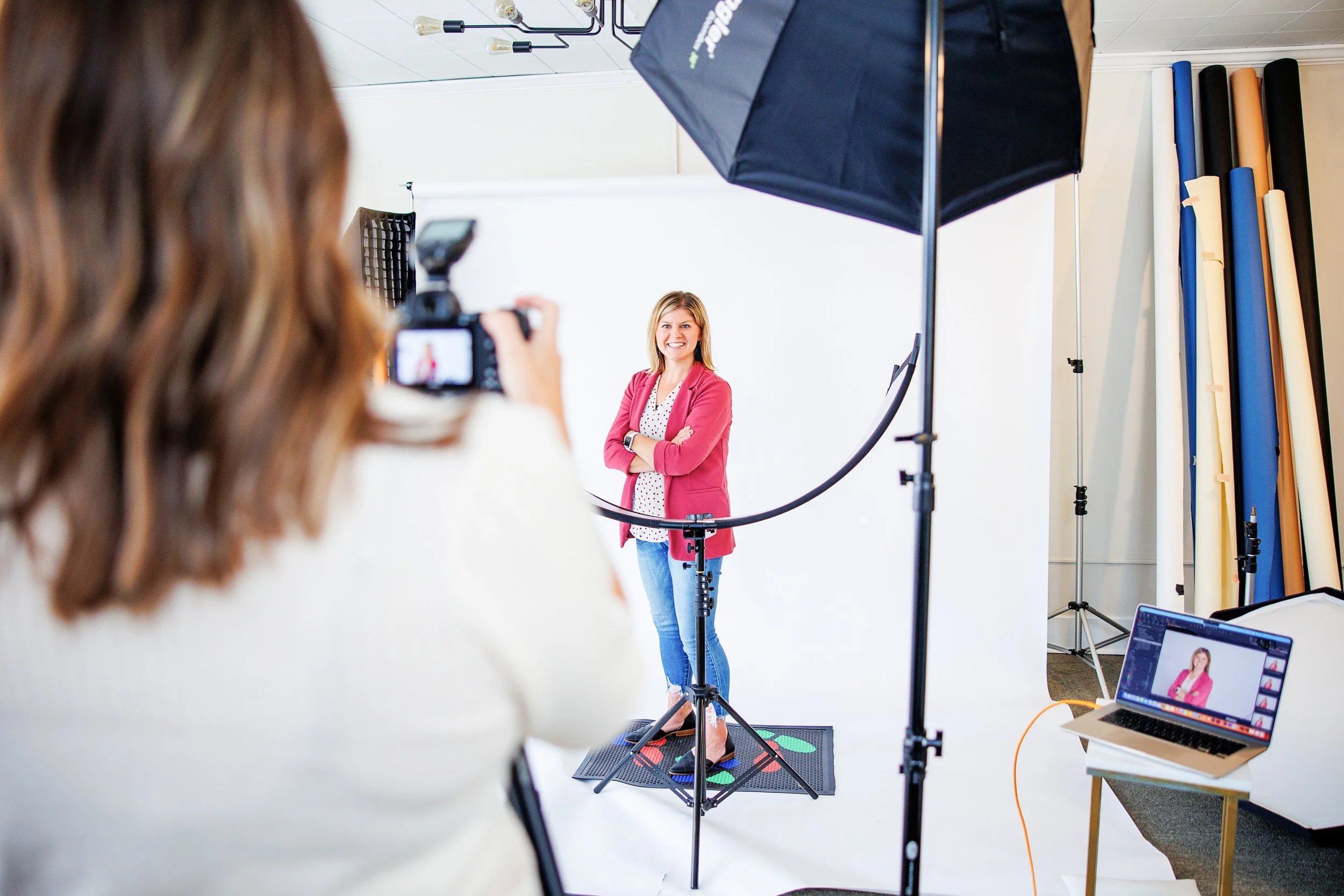 A woman in a pink blazer and jeans standing in a photography studio, smiling at the camera. She is on a photo light stand with a white backdrop behind her. A photographer is recording her with a camera, and a computer with a photo editing software is open on a table to the right.