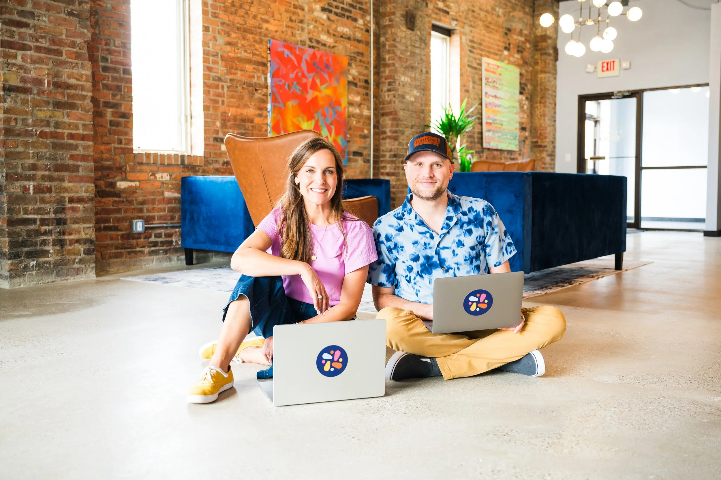 A woman and a man sitting on the floor with laptops in a modern, brightly lit office space with brick walls, colorful abstract artwork, a blue couch, and large windows.