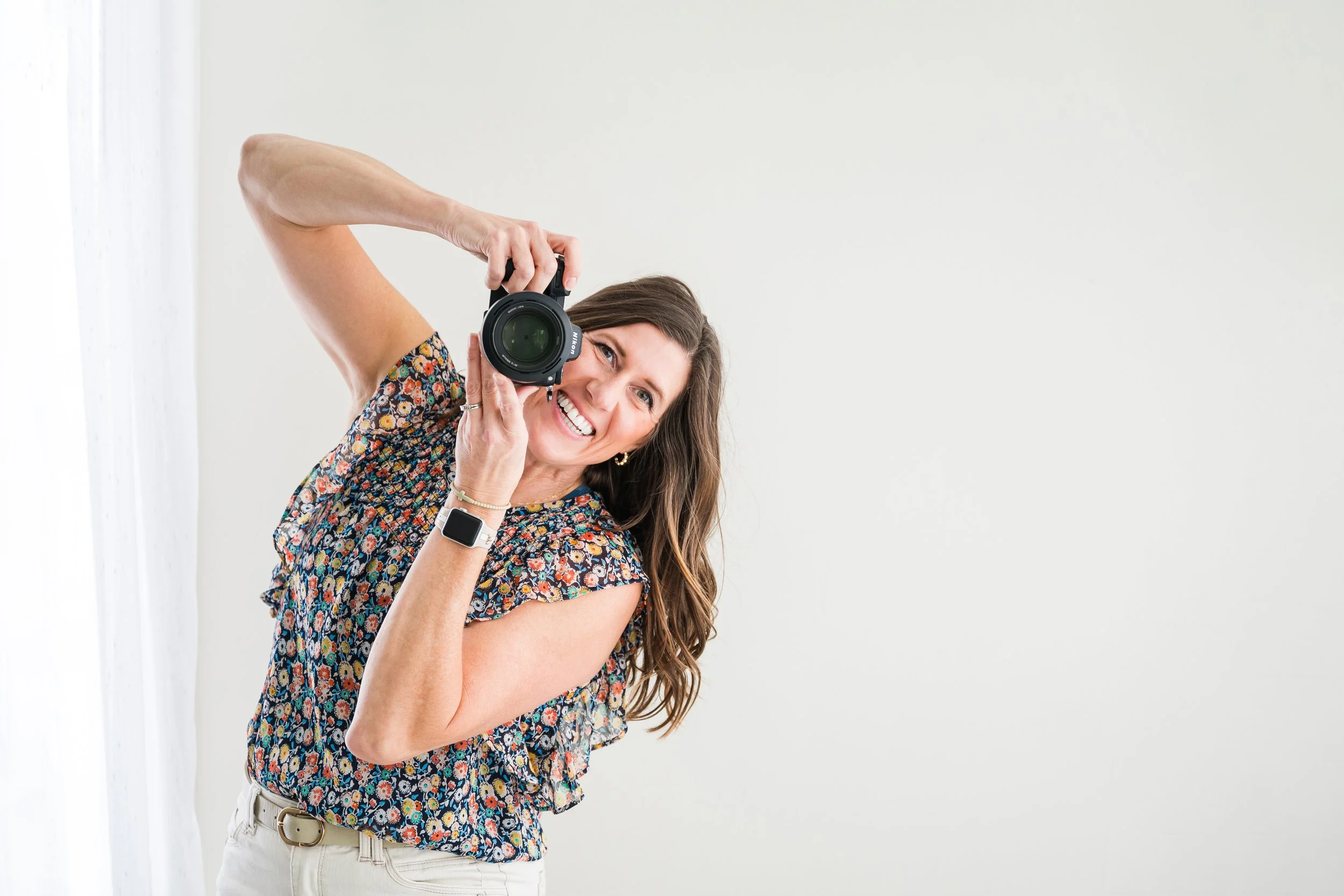 Woman with long brown hair, wearing a floral blouse and beige pants, holding a camera up to her face and smiling, standing against a plain white wall.