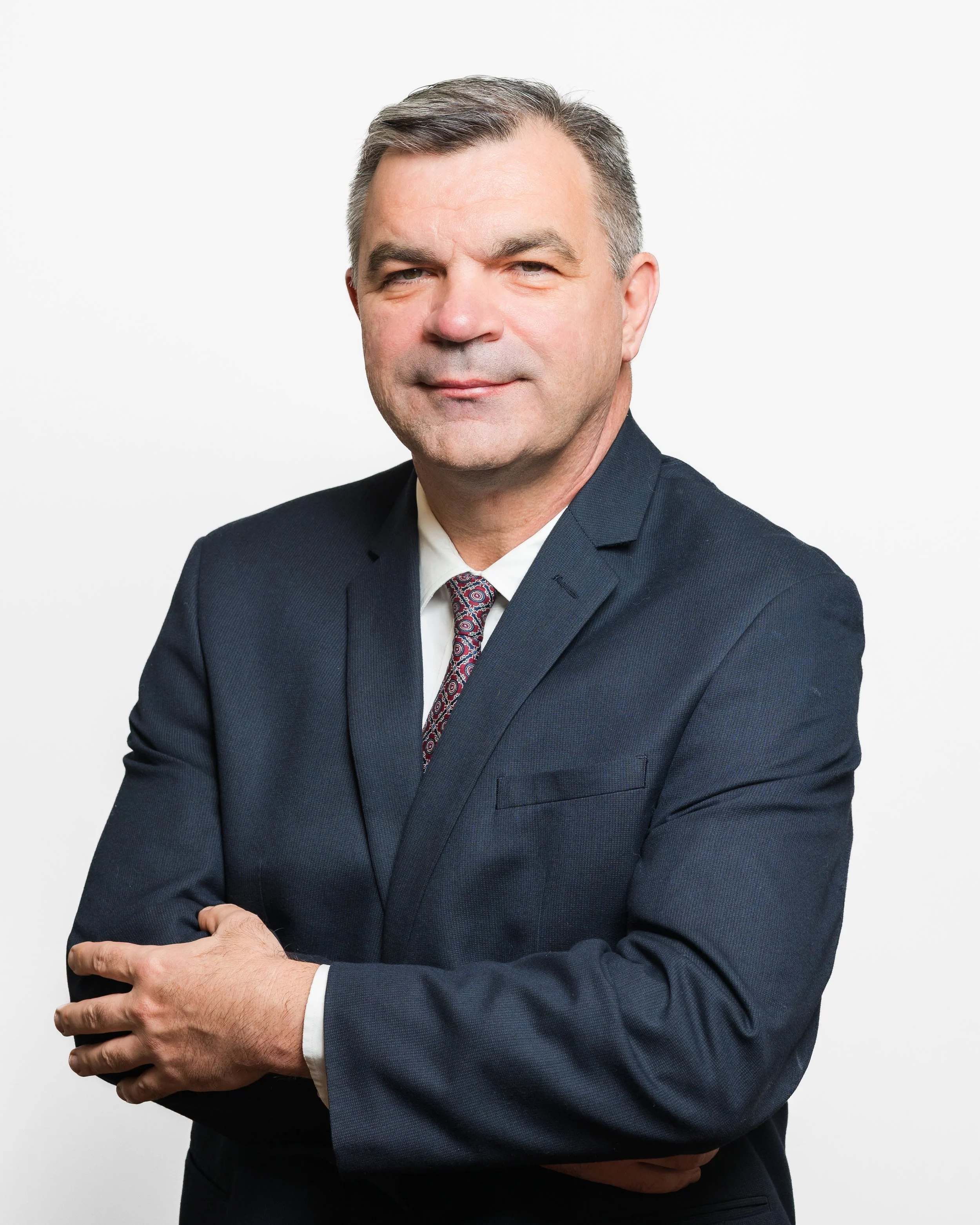 Professional portrait of a middle-aged man in a dark suit, white shirt, and patterned tie against a white background.