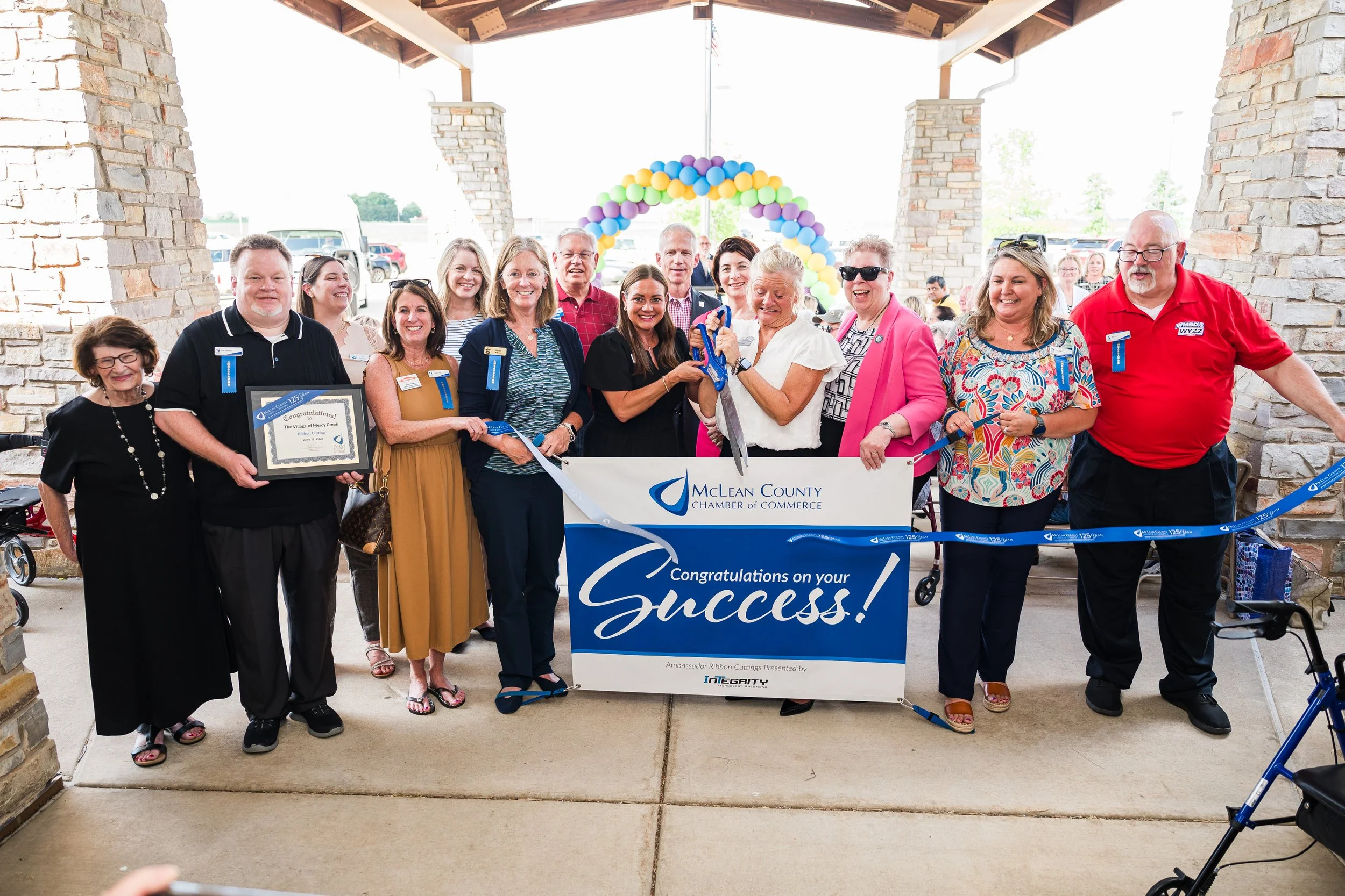 Group of people celebrating a ribbon-cutting event outdoors under a pavilion, with a congratulatory banner and colorful balloon arch in the background.