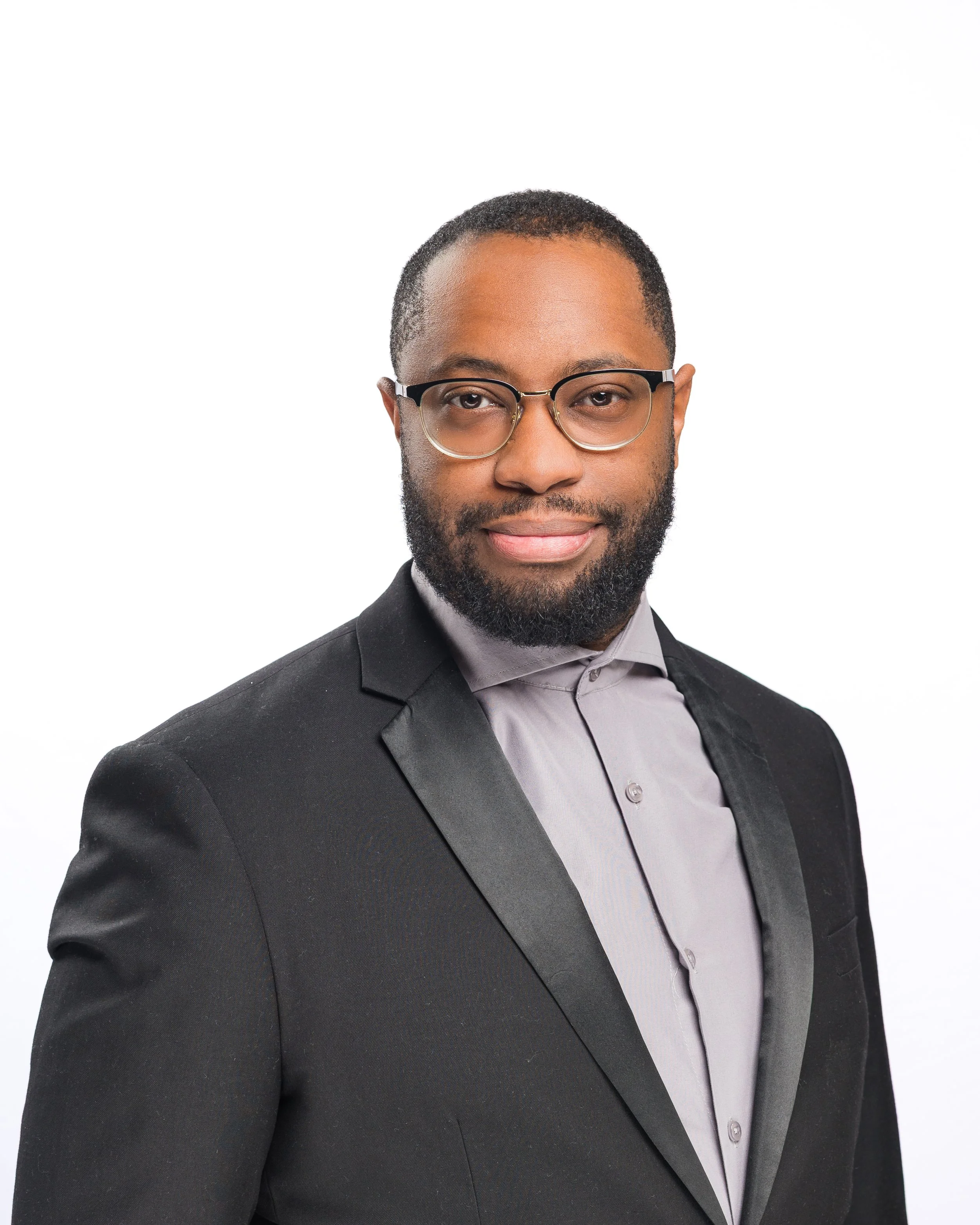 Professional headshot of a man with glasses, beard, wearing a black blazer and light gray shirt on a white background.